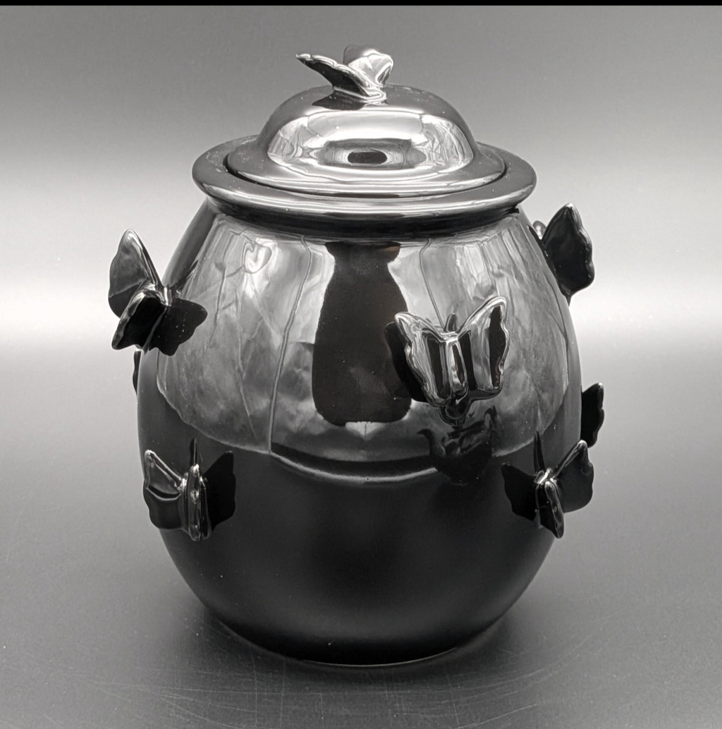 A glossy black ceramic jar with a matching lid, decorated with sculpted butterfly motifs on the sides and top, photographed against a neutral grey studio background.