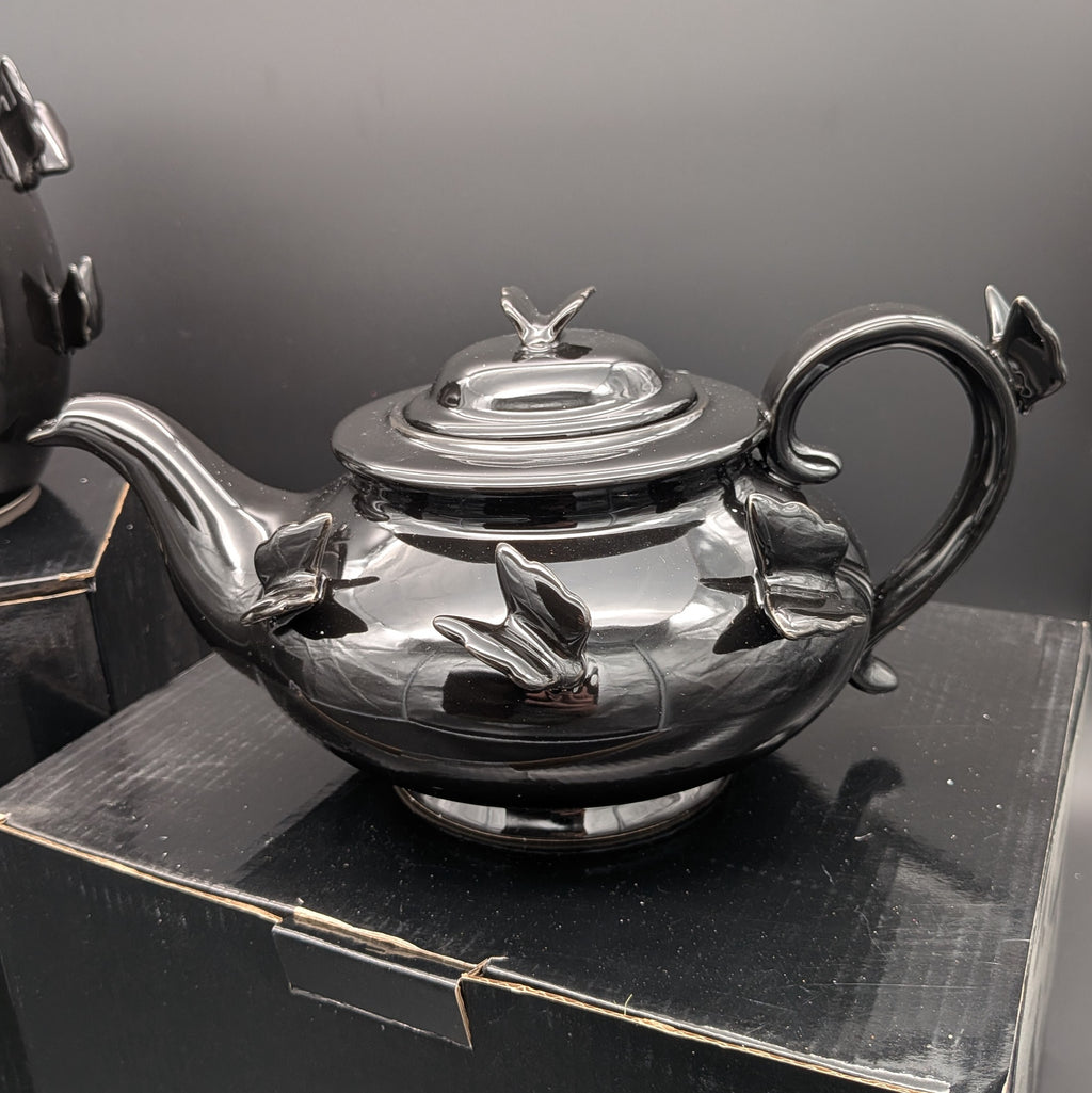 A polished black ceramic teapot with sculpted butterfly details on the body and lid, resting on a black display box against a dark grey background.