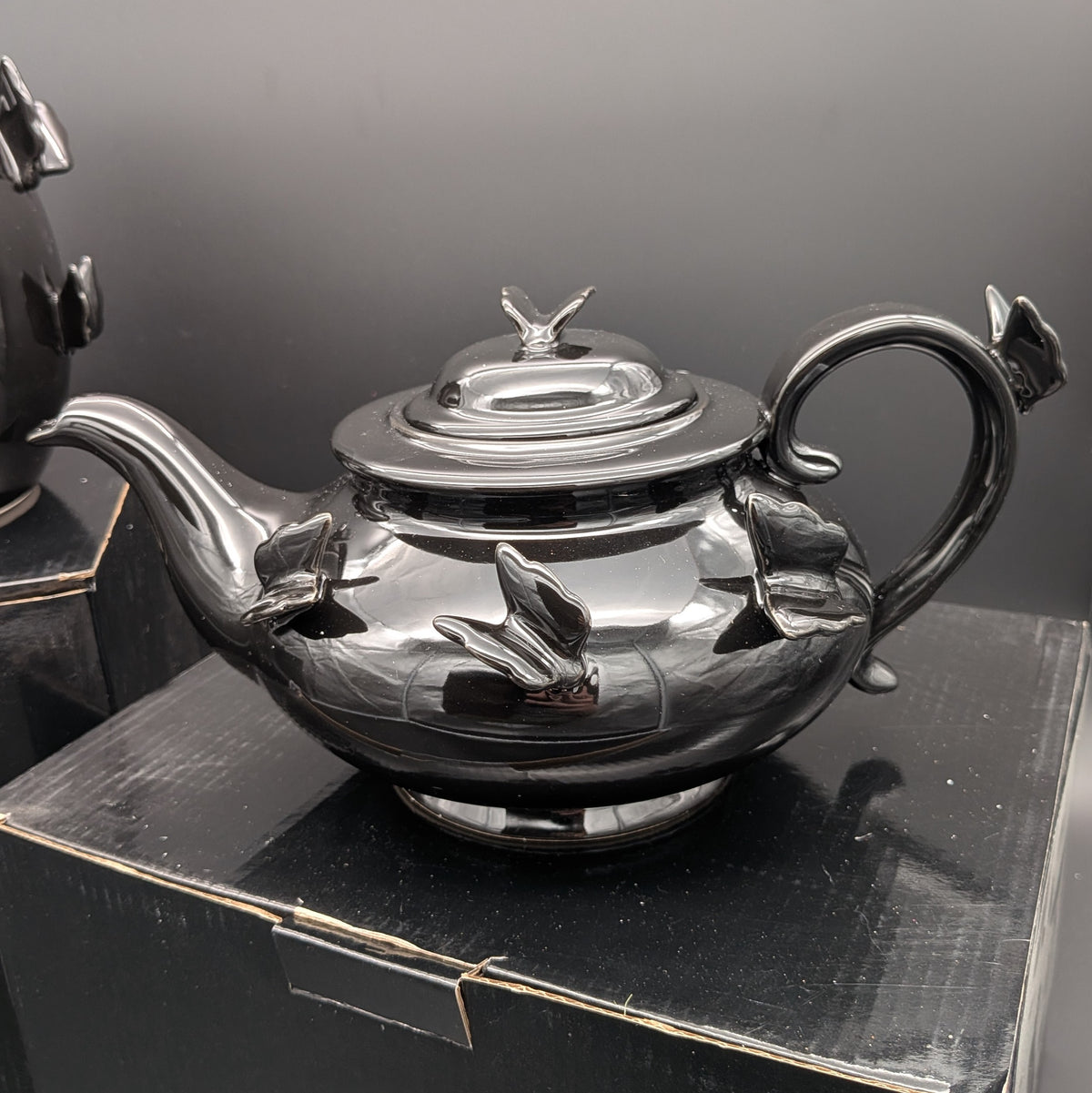 A polished black ceramic teapot with sculpted butterfly details on the body and lid, resting on a black display box against a dark grey background.