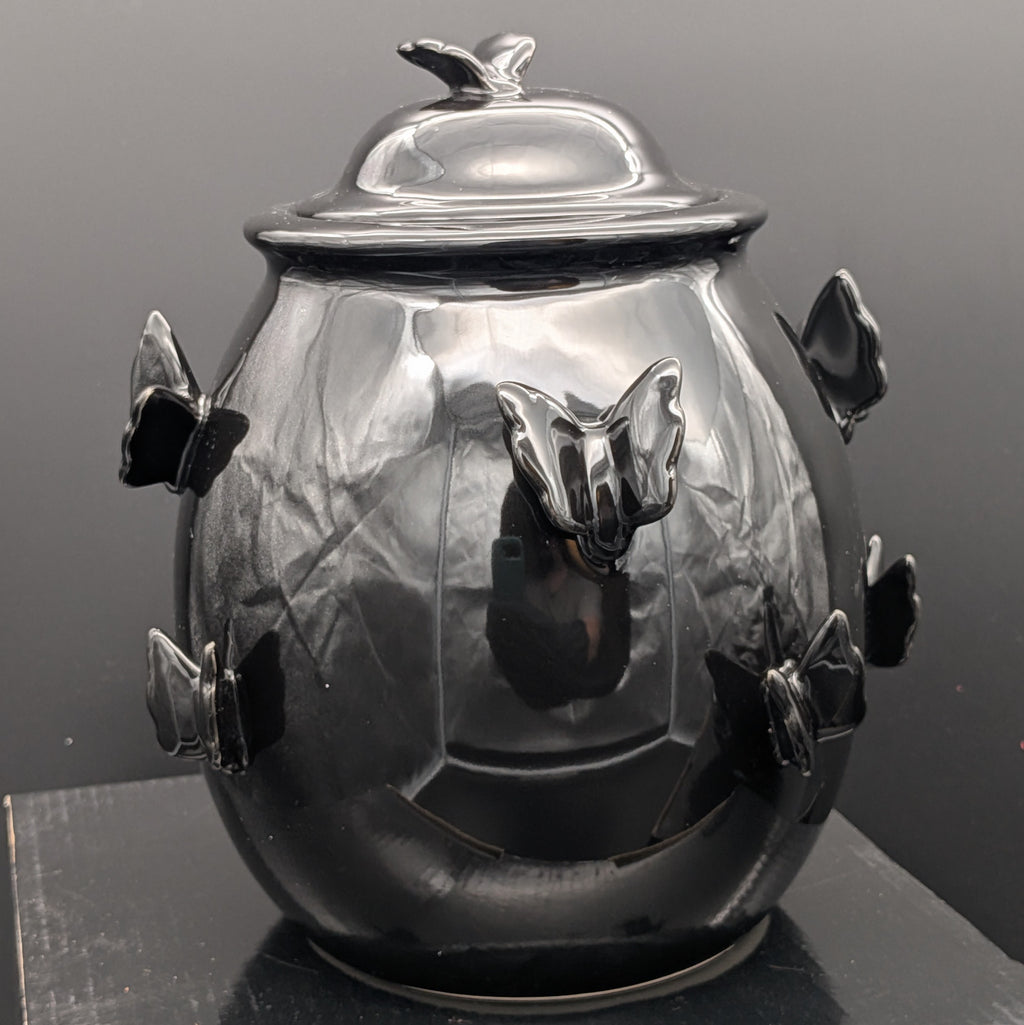 A gothic, glossy black ceramic jar with a lid, decorated with three dimensional black butterfly motifs on the front and sides, set against a dark grey background. From Haunted Homeware by Killstar