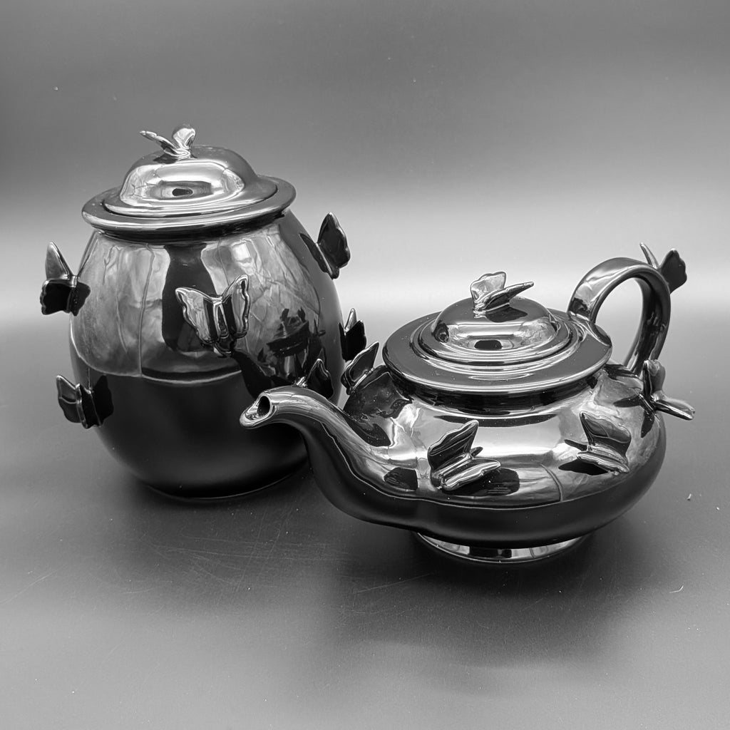 A gothic matching teapot and jar set, in polished gloss black finish with sculpted  butterfly details, photographed on a dark grey studio background
