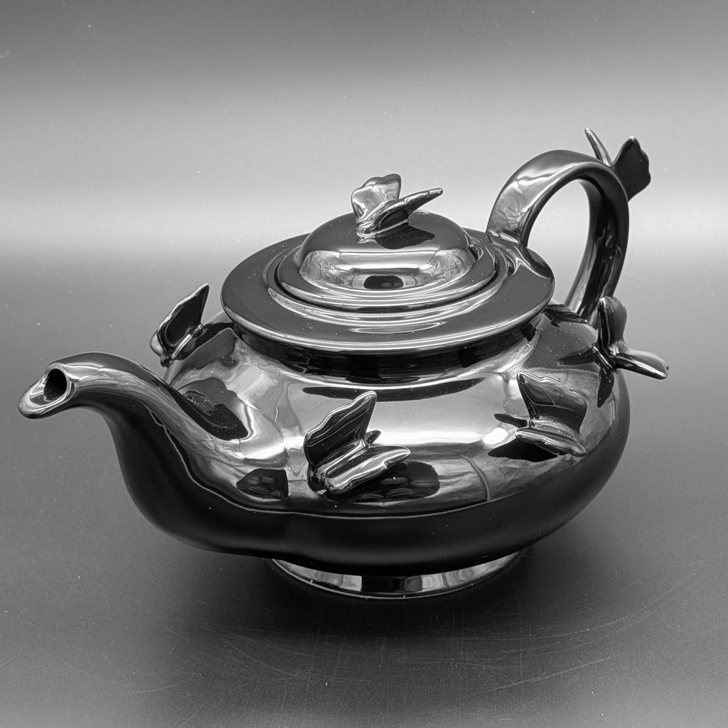 A highly polished black ceramic teapot with sculpted butterfly motifs on the body, handle and lid topper, photographed on a smooth grey studio background. A beautiful item from Killstar.