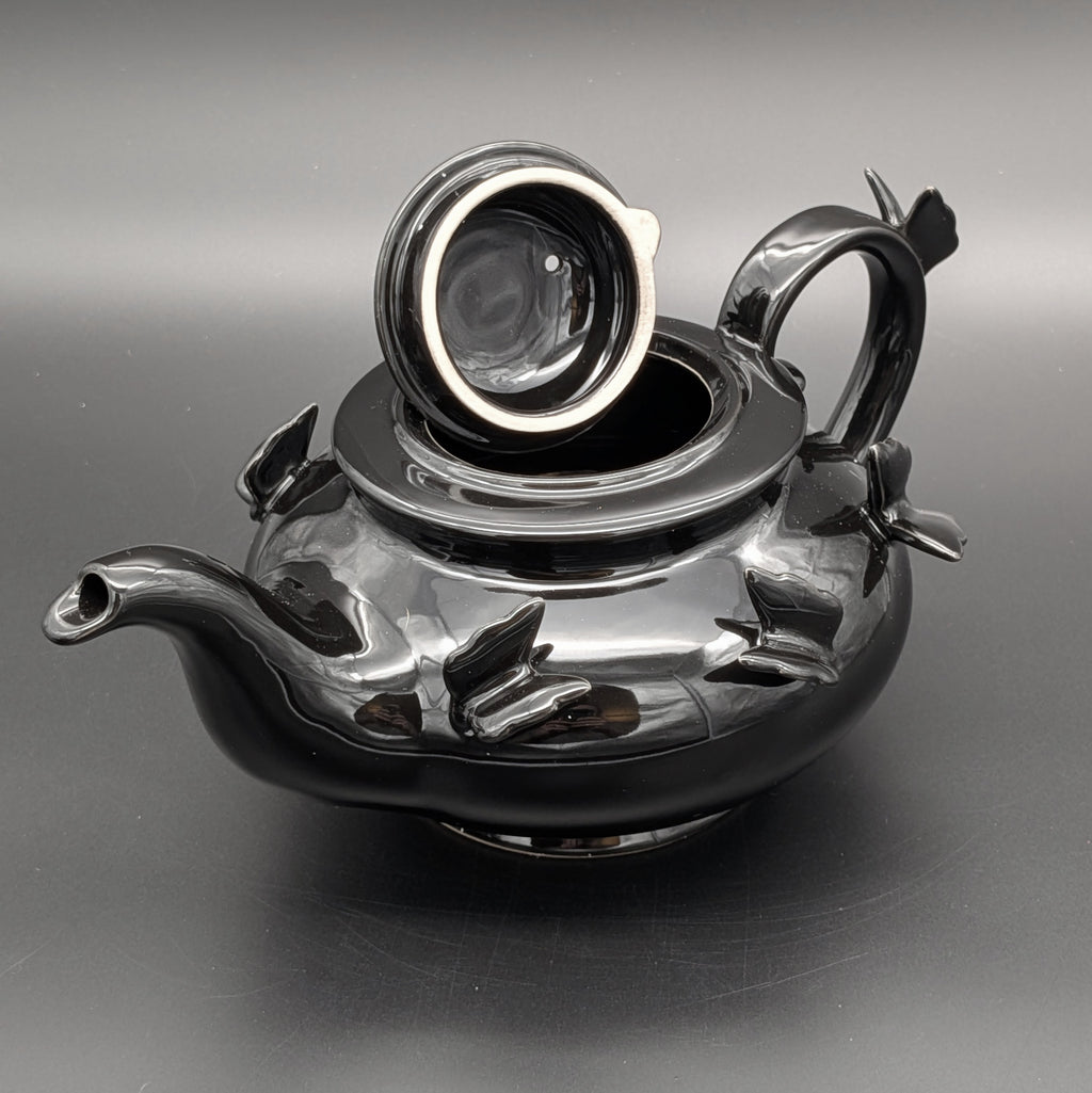 A glossy black ceramic teapot with sculpted butterfly details on the body &  matching lid, set against a dark gray background. Gothic fairy inspired