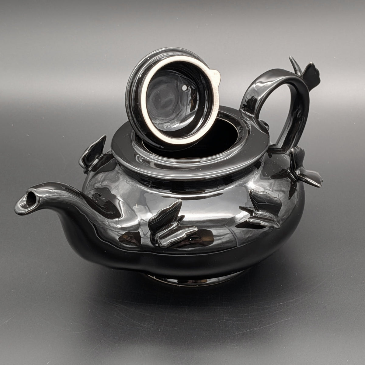 A glossy black ceramic teapot with sculpted butterfly details on the body &  matching lid, set against a dark gray background. Gothic fairy inspired
