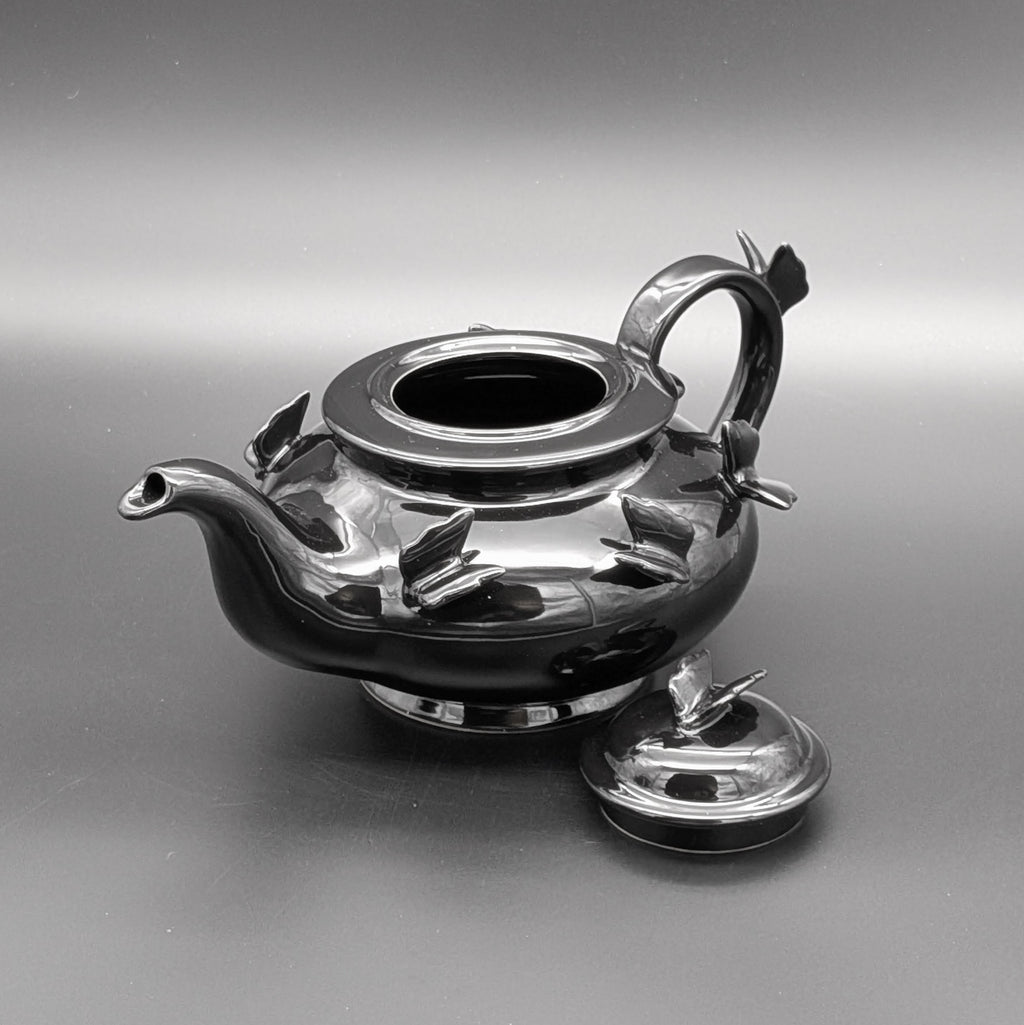 A glossy black teapot with sculpted butterfly details on its body, a matching lid resting beside it, and a reflective grey gradient studio background.