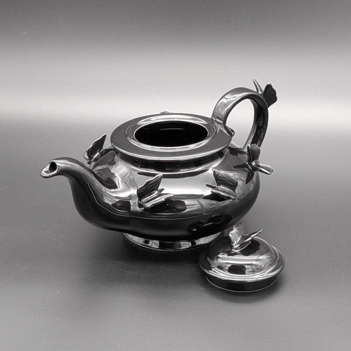A glossy black teapot with sculpted butterfly details on its body, a matching lid resting beside it, and a reflective grey gradient studio background.