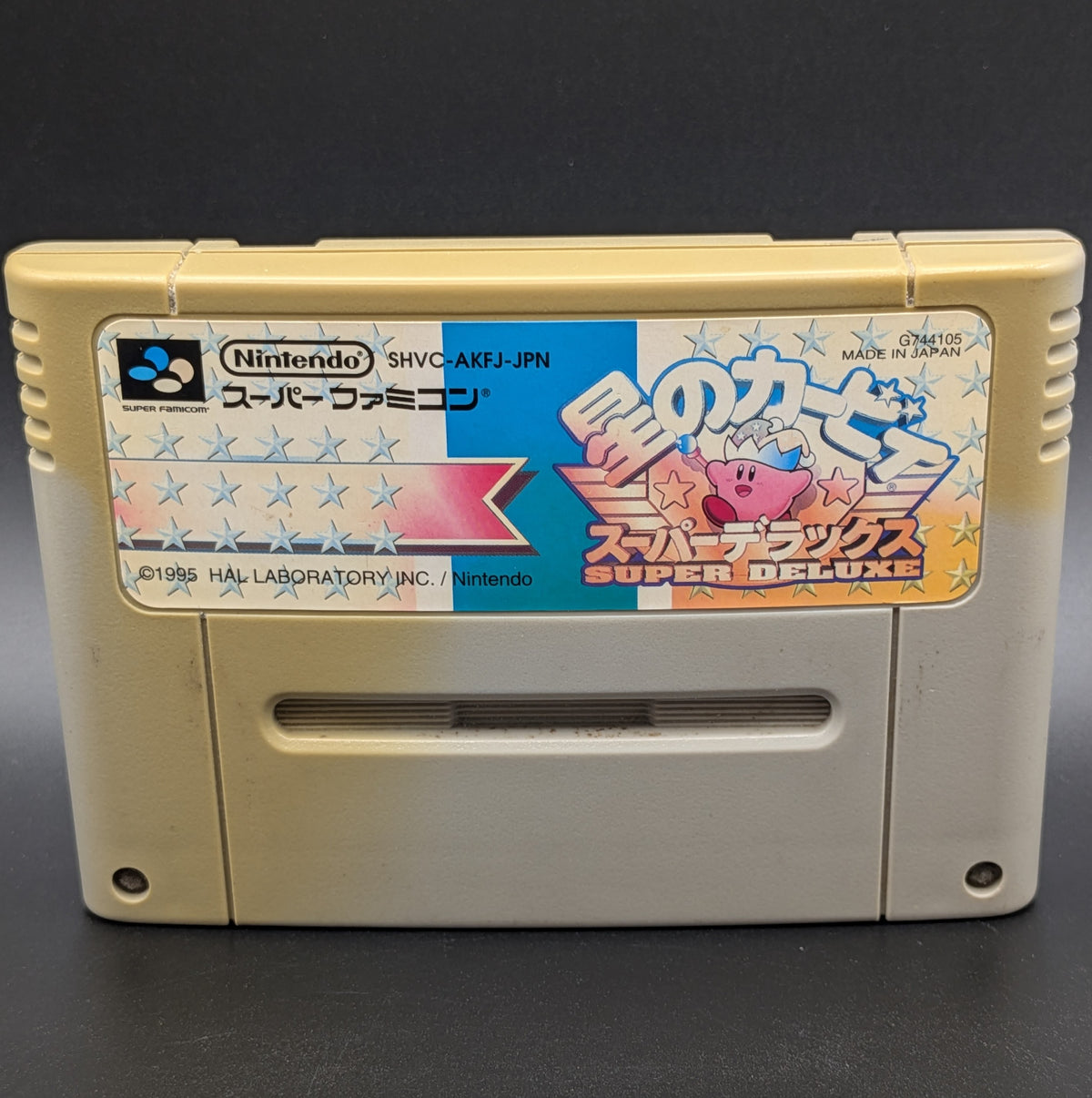 A close up of Kirby's Dream Land Super Deluxe video game cartridge for Nintendo's Super Famicom (SFC) Entertainment System, shown standing up and facing forwards. It has large areas of yellowing and the label has faded. Displayed a dark grey background.
