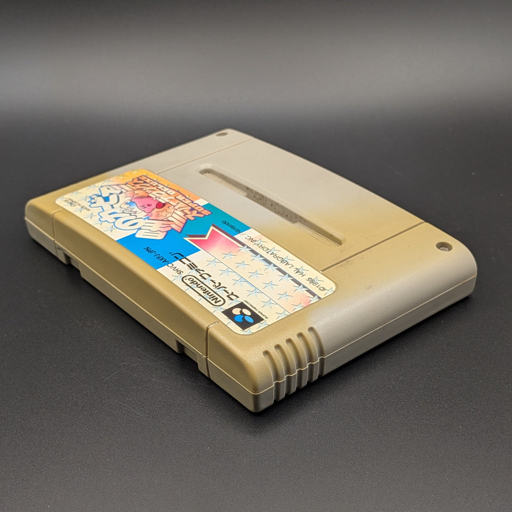 Kirby's Dream Land Super Deluxe video game cartridge for Nintendo's Super Famicom (SFC) Entertainment System, shown laying on its back with the label facing upwards. Showing the left hand side and top edge. It has large areas of yellowing and the label has faded. Displayed a dark grey background.