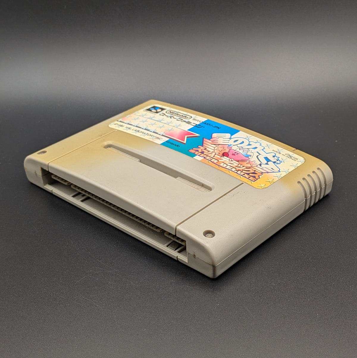 Kirby's Dream Land Super Deluxe video game cartridge for Nintendo's Super Famicom (SFC) Entertainment System, shown laying on its back with the label facing upwards. Showing the right hand side and bottom slot. It has large areas of yellowing and the label has faded. Displayed a dark grey background.