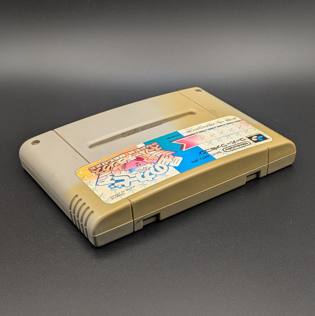 Kirby's Dream Land Super Deluxe video game cartridge for Nintendo's Super Famicom (SFC) Entertainment System, shown laying on its back with the label facing upwards. Showing the right hand side and top edge. It has large areas of yellowing and the label has faded. Displayed a dark grey background.
