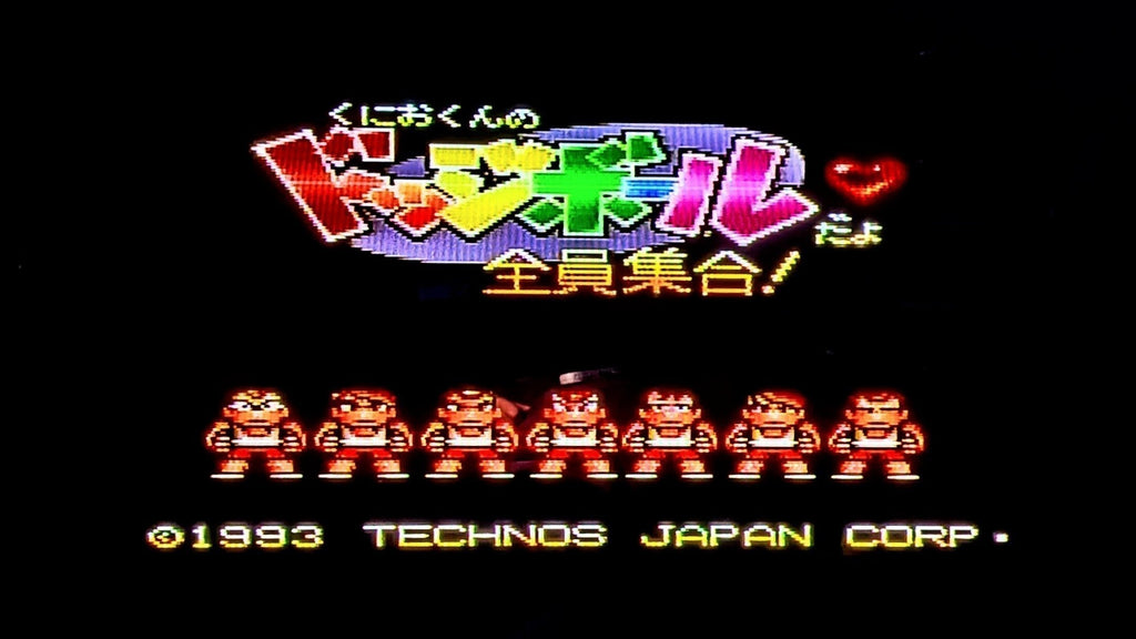 The game title screen for Nintendo's Kunio-Kun no Dodgeball for the Super Famicom (SFC), featuring colourful block Japanese text sitting over a purple striped oval. Underneath are 7 characters in a line, facing the screen in white tops and red shorts, at the bottom it reads (C)1993 TECHNOS JAPAN CORP. All on a black background. This show sthe game is tested and working.