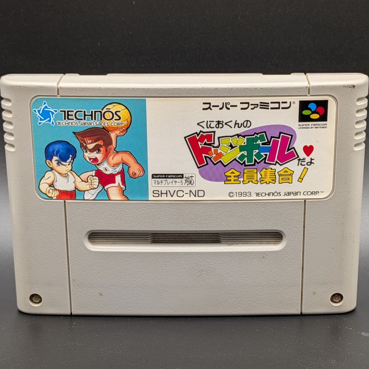 A close up of Kunio-Kun no Dodgeball video game cartridge for Nintendo's Super Famicom (SFC) Entertainment System, shown standing up and facing forward on a dark grey background.
