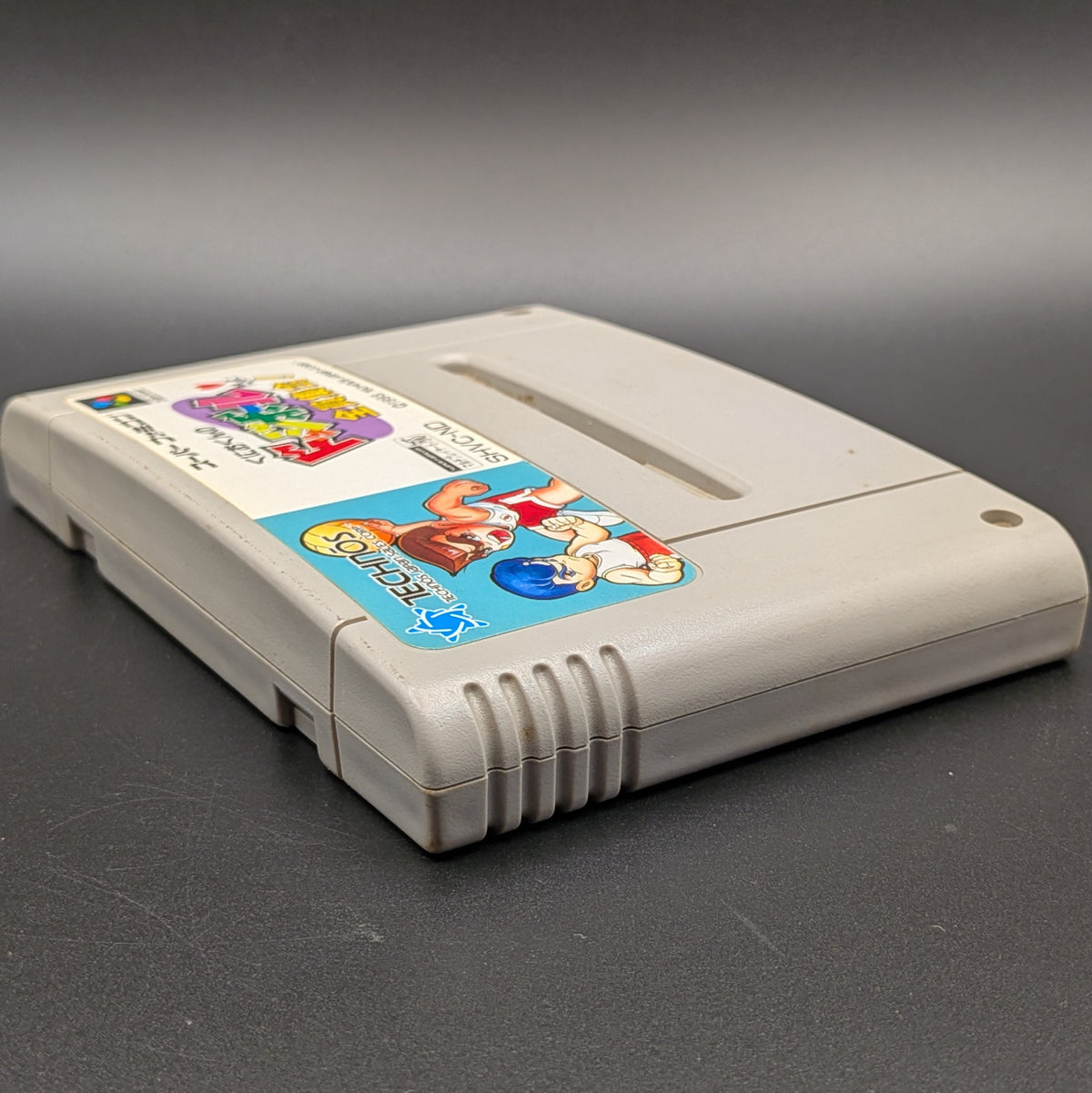 Kunio-Kun no Dodgeball video game cartridge for Nintendo's Super Famicom (SFC) Entertainment System, shown laying on its back with the label facing upwards. Showing the left hand side and top edge. It sits on a dark grey background