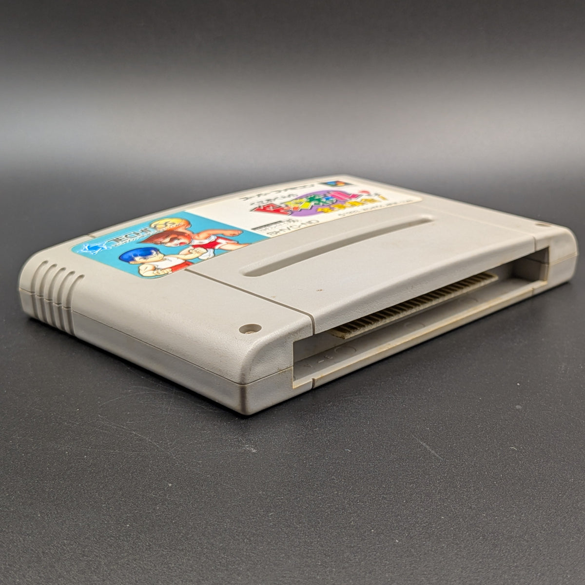 Kunio-Kun no Dodgeball video game cartridge for Nintendo's Super Famicom (SFC) Entertainment System, shown laying on its back with the label facing upwards. Showing the left hand side and bottom slot. It sits on a dark grey background
