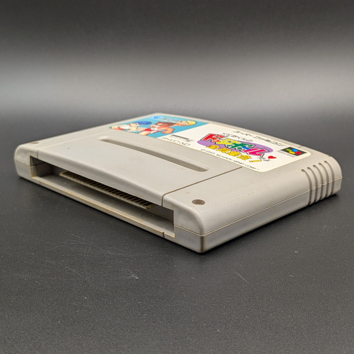 Kunio-Kun no Dodgeball video game cartridge for Nintendo's Super Famicom (SFC) Entertainment System, shown laying on its back with the label facing upwards. Showing the right hand side and bottom slot. It sits on a dark grey background