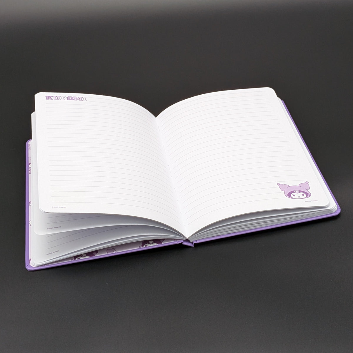 A purple Kuromi ruled notebook laying open revealing the lined paper and Kuromi motiffs in the corners, photographed on a dark gray background. From the Sanrio POP range, printed by Blue Sky Studios.