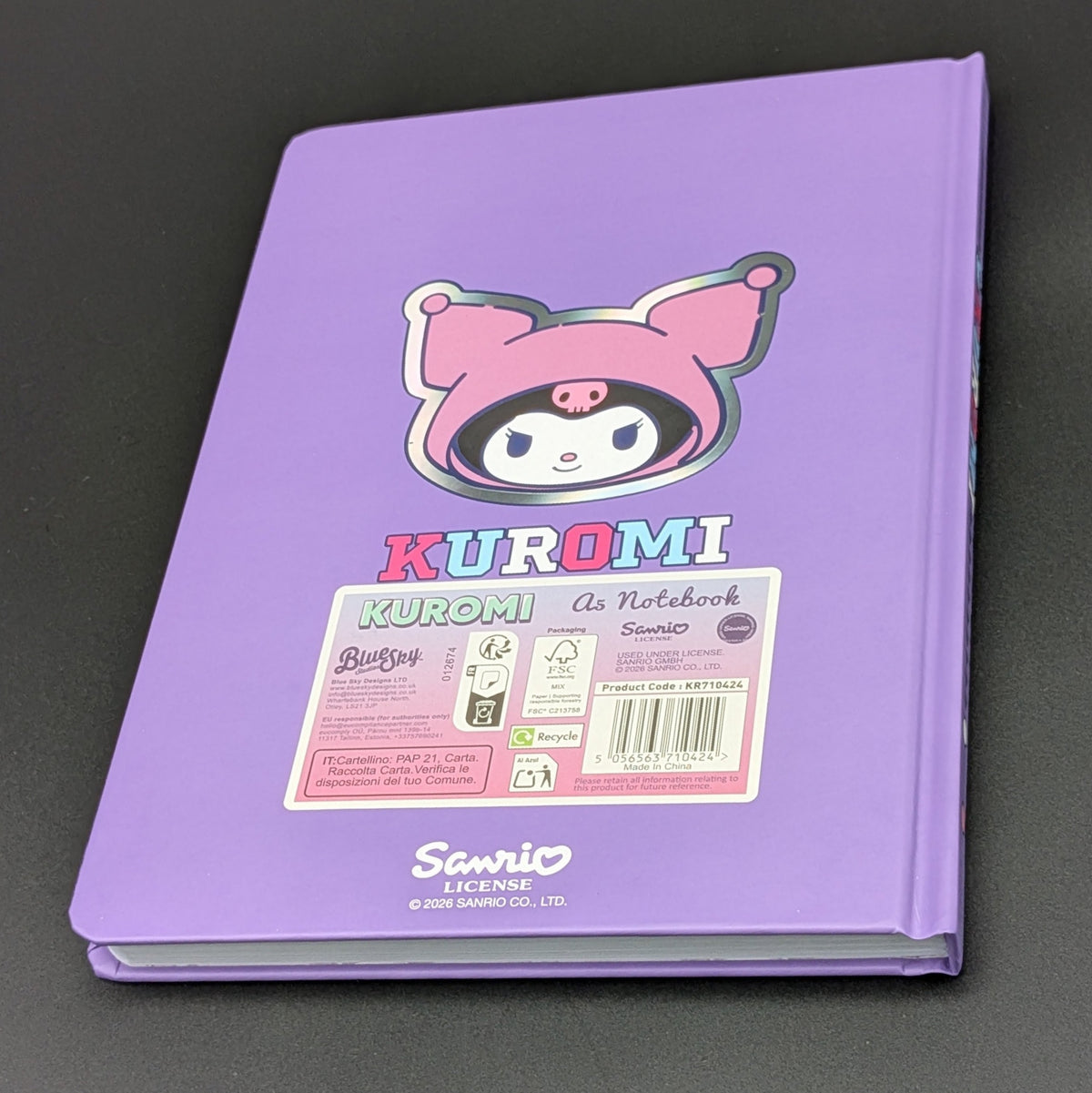 A purple Kuromi ruled notebook with a colourful cartoon illustration on the back and a label with product information, photographed on a dark gray background. From the Sanrio POP range, printed by Blue Sky Studios.