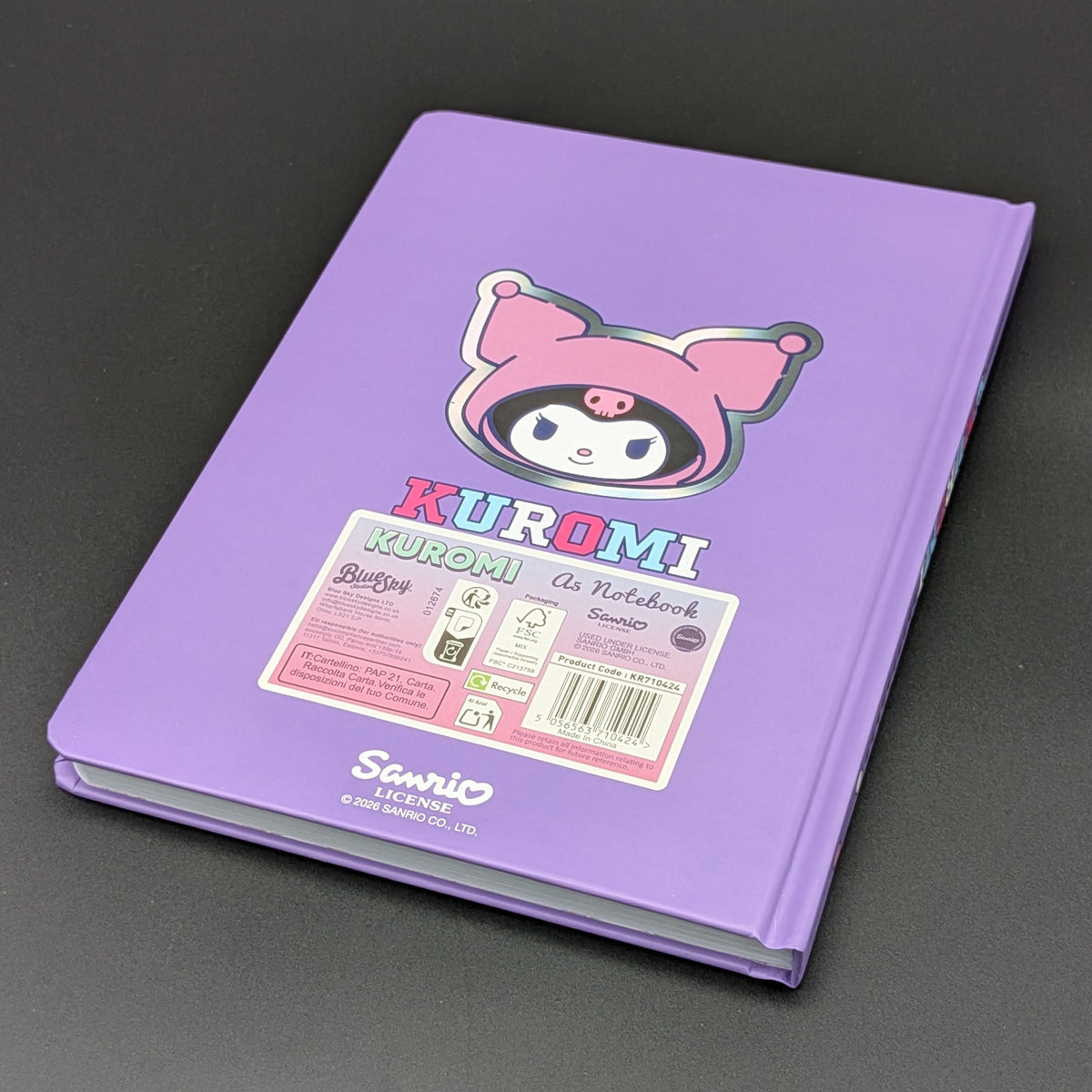 A purple Kuromi ruled notebook with a colourful cartoon illustration on the back and a label with product information, sitting t a 3/4 view angle & photographed on a dark gray background. From the Sanrio POP range, printed by Blue Sky Studios.