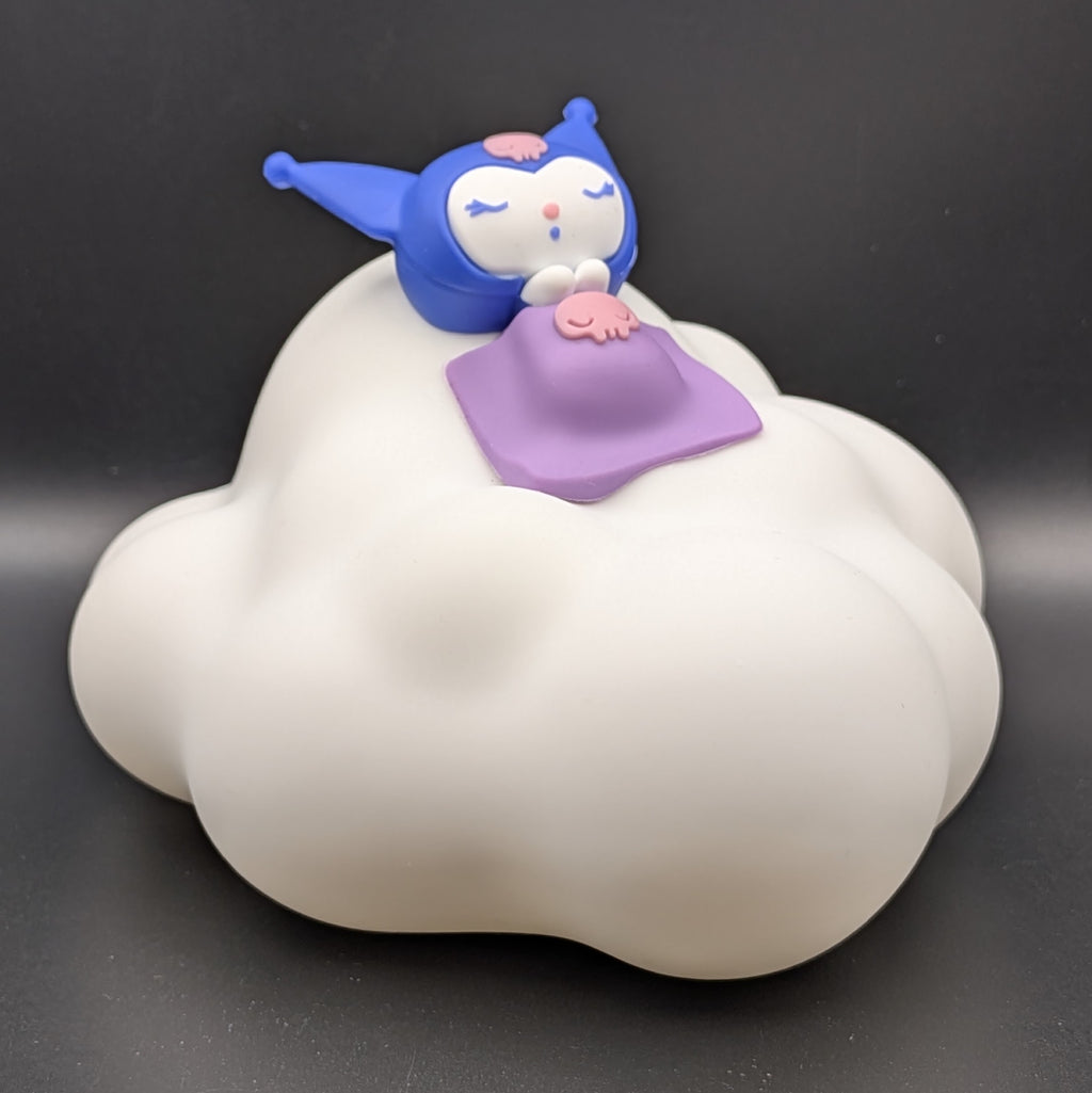 A small Kuromi figurine sleeping on a white cloud-shaped base, with a purple blanket and pink details, photographed against a dark gray background.