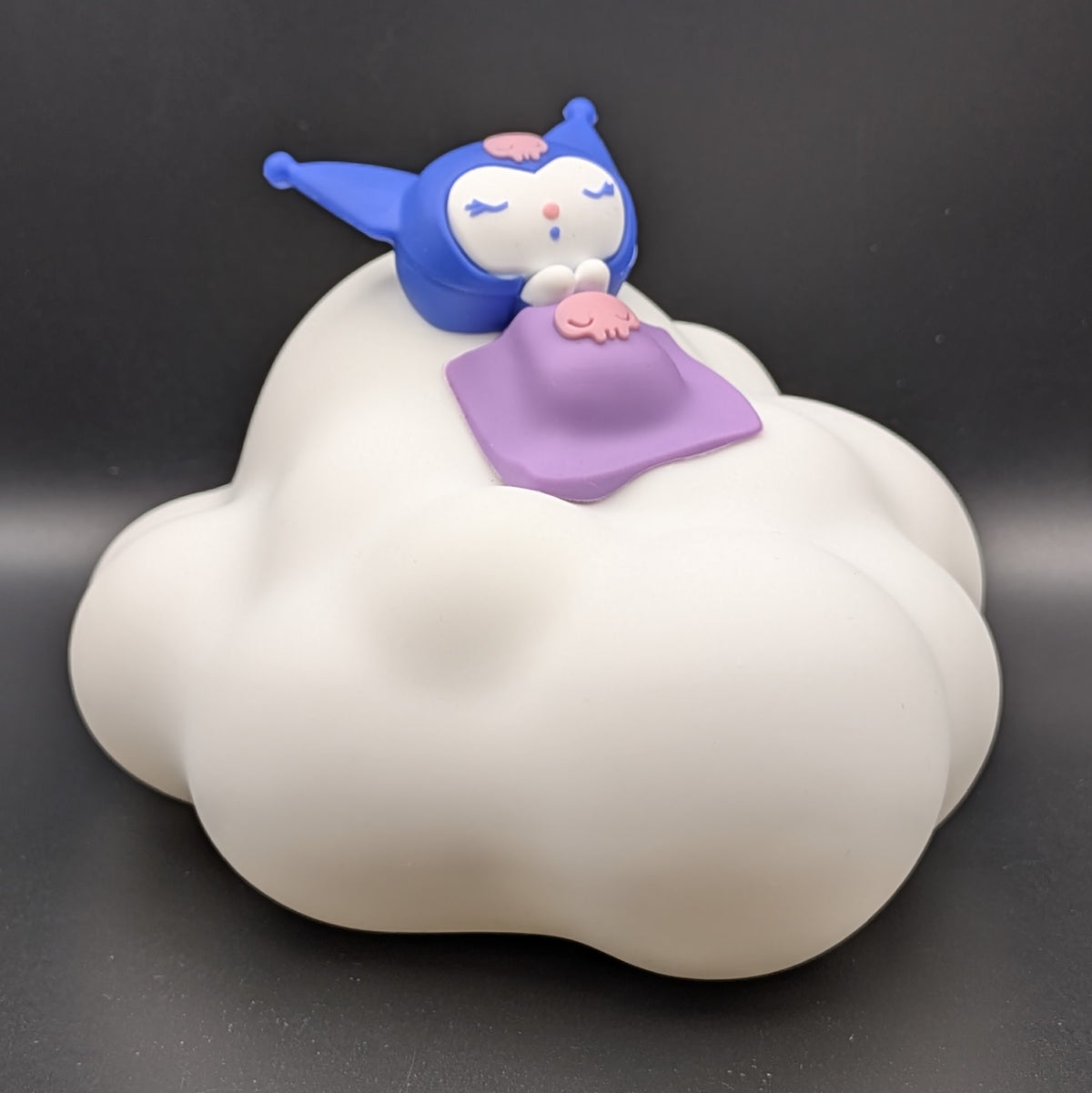 A small Kuromi figurine sleeping on a white cloud-shaped base, with a purple blanket and pink details, photographed against a dark gray background.