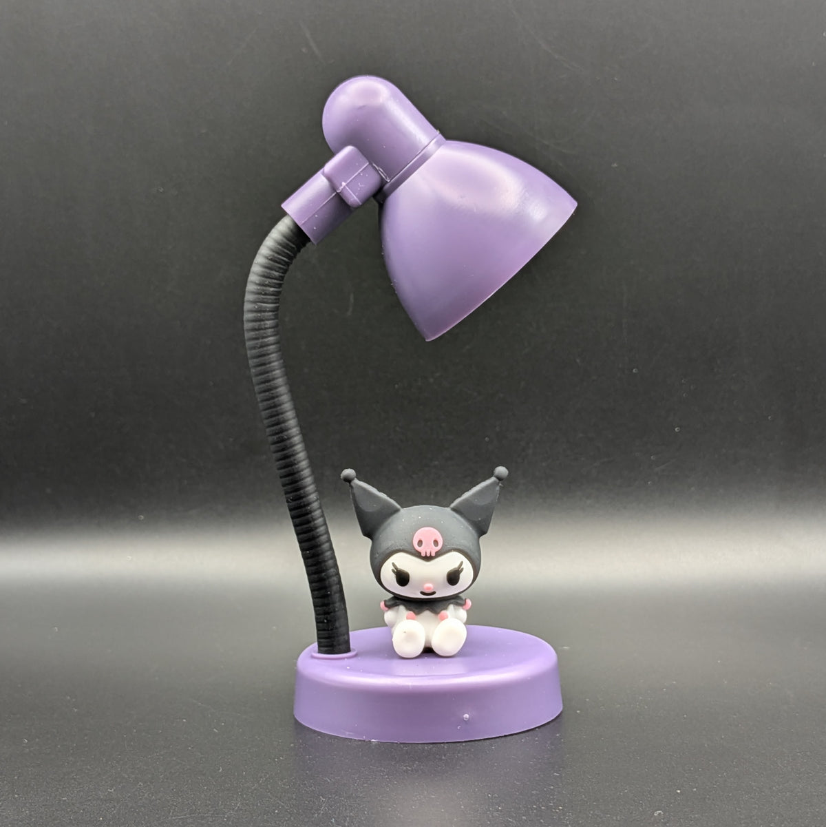 A small tabletop lamp with a purple dome-shaped shade, black flexible arm, and round purple base featuring a cute dark-gray character figurine with white paws and a pink forehead mark, photographed against a dark gray studio background.