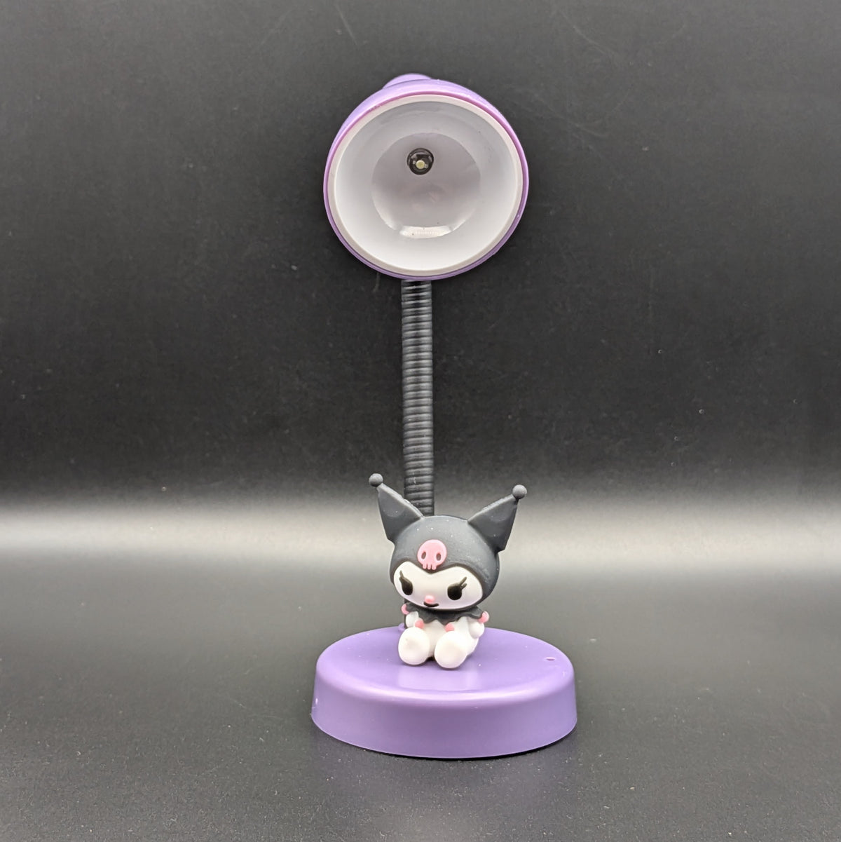 A small tabletop lamp featuring a cartoon character figure seated on a round purple base, with a flexible black arm and a purple adjustable head, photographed against a dark gray background.
