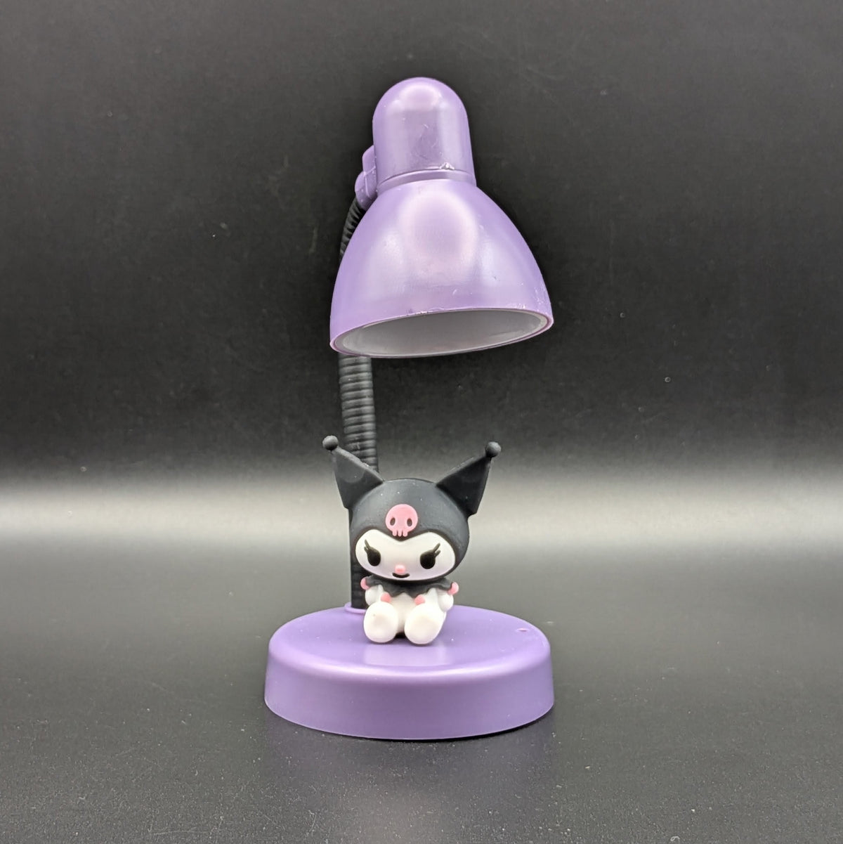 A small tabletop lamp featuring a purple dome-shaped shade, a black flexible arm, and a round purple base with a cute black-and-white character figurine sitting on it, photographed against a dark gray background.