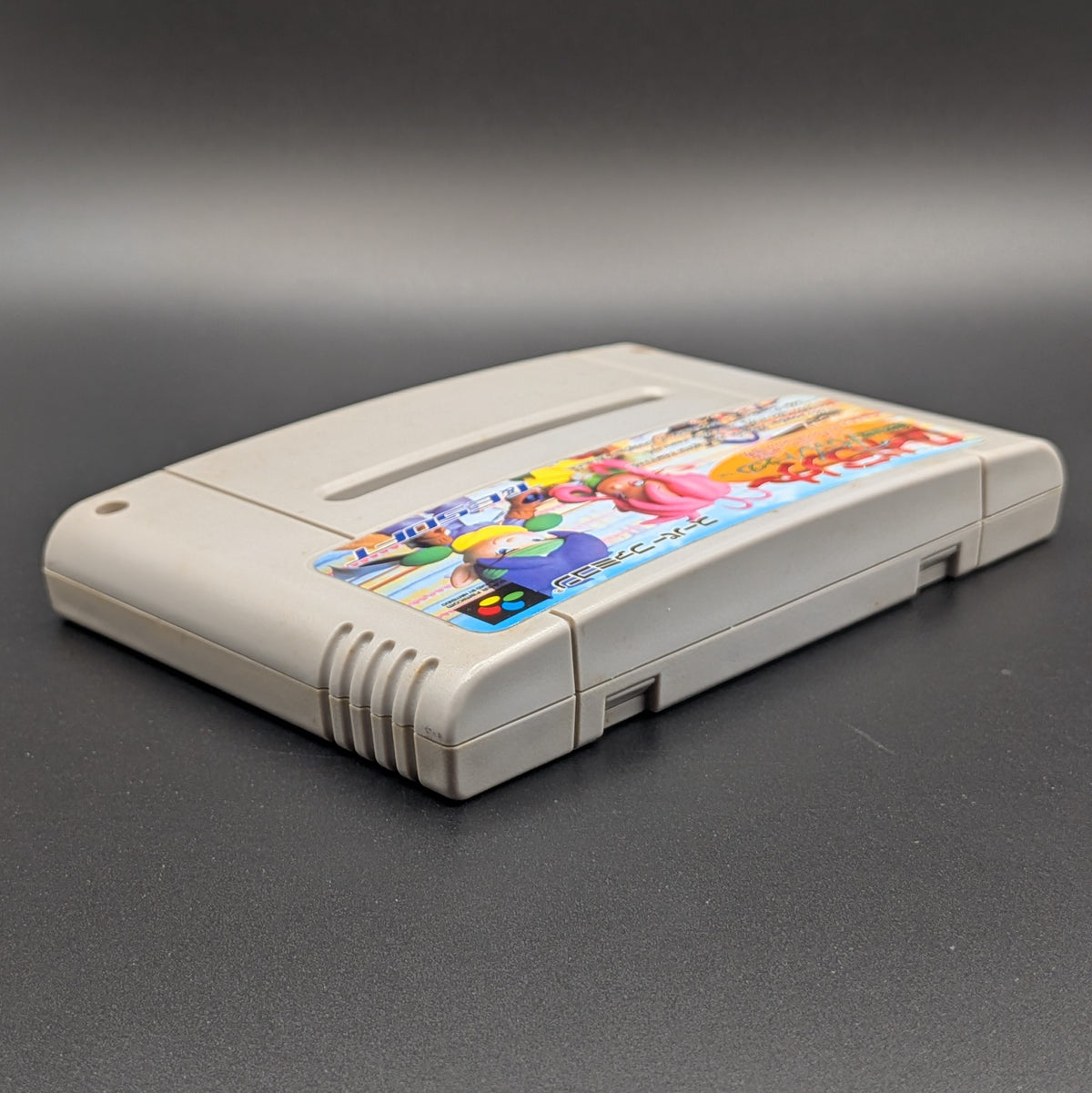 Lode Runner Twin : Justy to Liberty Daibouken video game cartridge for Nintendo's Super Famicom (SFC) Entertainment System, shown laying on its back with the label facing upwards. Showing the right hand side and top edge. It sits on a dark grey background. 