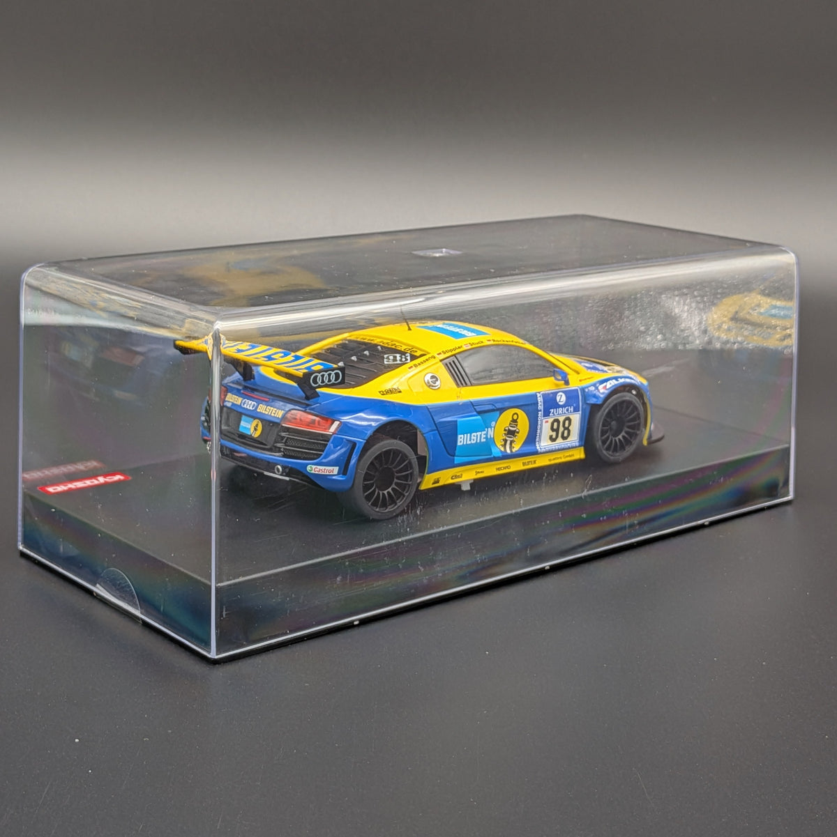 A diecast model of a blue and yellow Audi R8 race car inside a clear plastic display case, photographed on a dark gray background.