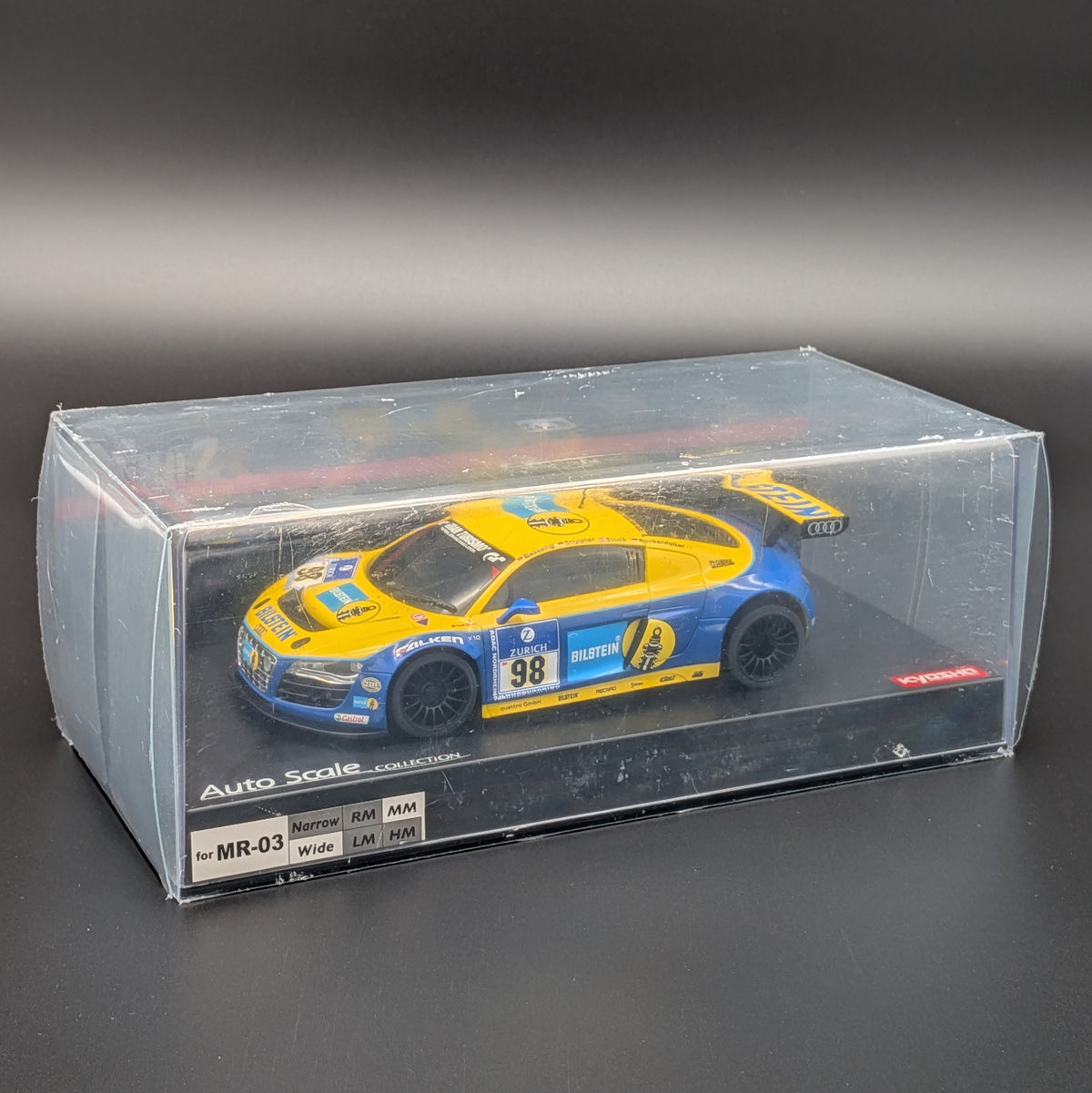 A 1:27 scale model car of a blue and yellow Audi R8 Phoenix Racing LMS race car, displayed inside its clear plastic display box on a dark gray background.