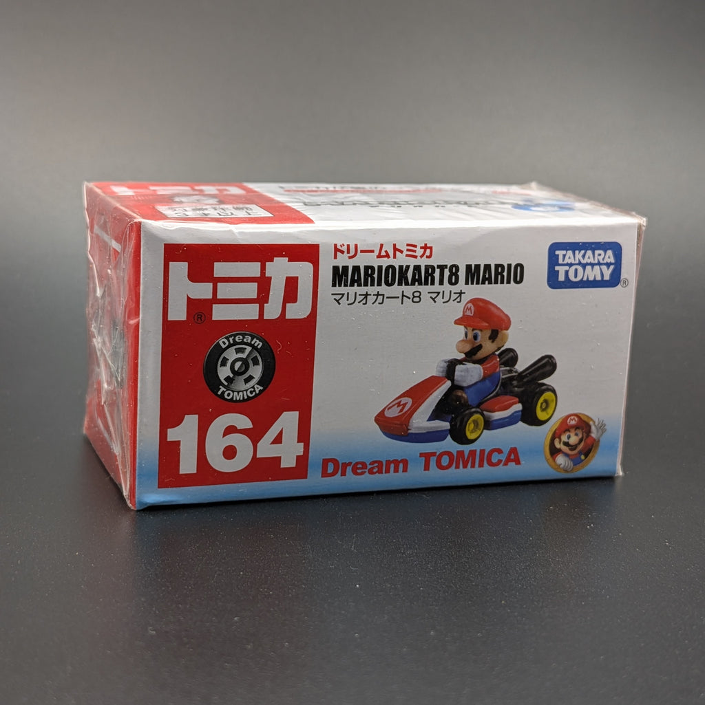 A boxed Dream TOMICA Mario Kart 8 Mario toy car package, shown on a dark gray surface. The box is white with red and blue sections, large white '164' text, and a printed image of Mario in a red and blue race kart.