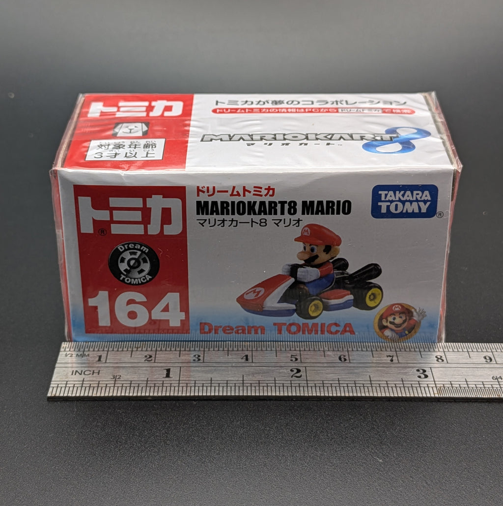 A boxed Dream TOMICA Mario Kart 8 Mario toy car package standing upright on a dark surface, with a metal ruler placed in front for scale. The box is white with red and blue sections and printed product imagery including a red race car and Takara Tomy branding.