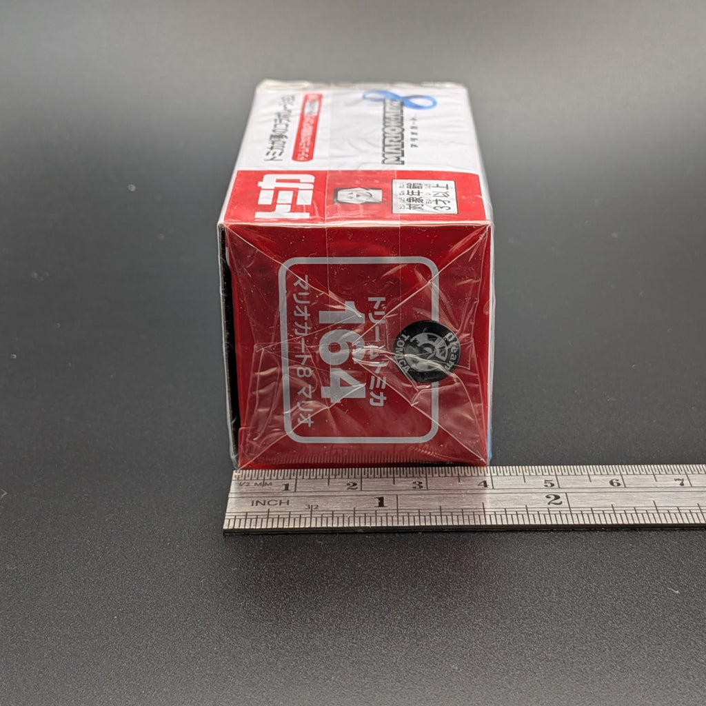 A red and white box of 1/6 scale model kits, specifically Tomica 164, shown with a metal ruler on a dark gray surface to indicate its size.