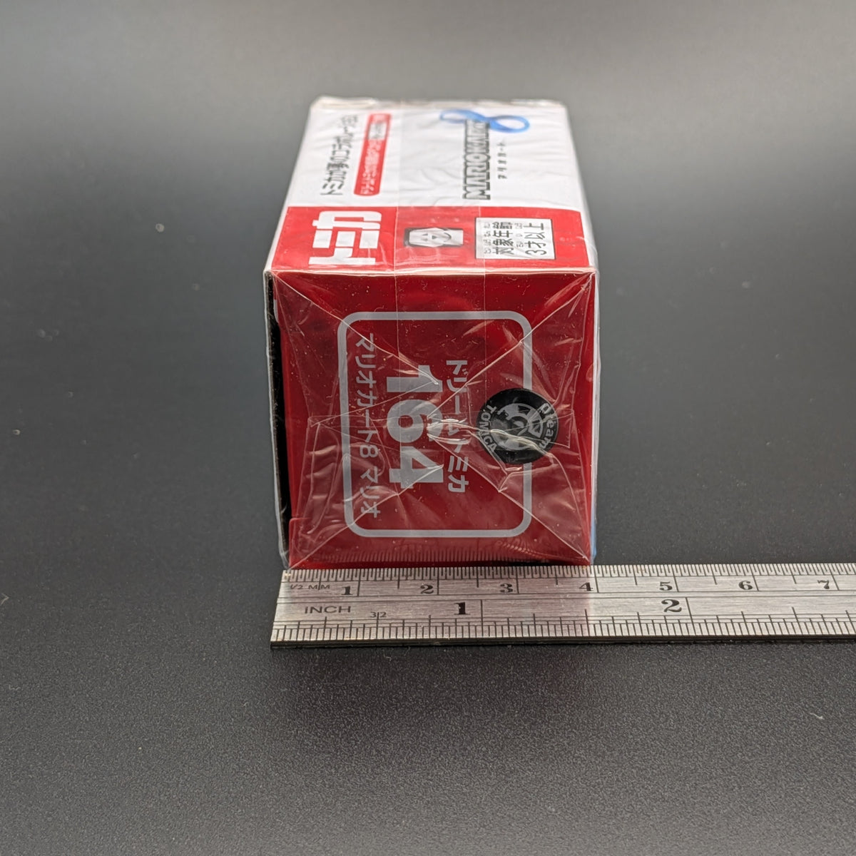 A red and white box of 1/6 scale model kits, specifically Tomica 164, shown with a metal ruler on a dark gray surface to indicate its size.