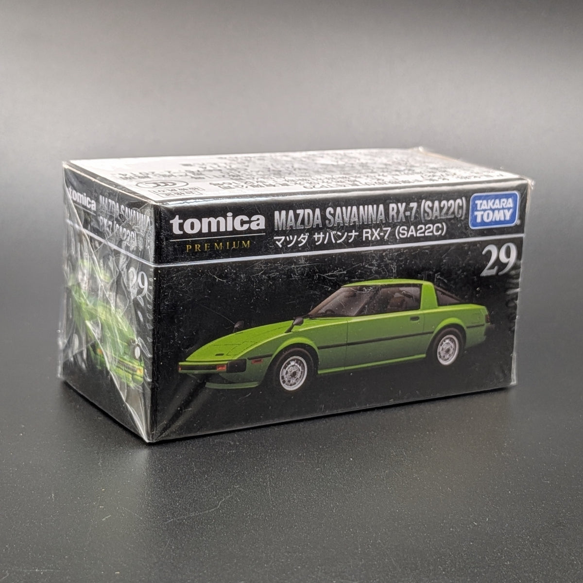 A boxed Tomica Mazda Savanna RX-7 model car, still wrapped in clear plastic, on a dark gray background. The box shows a green sports car and the text 'tomica', 'MAZDA SAVANNA RX-7 (SA22C)', and 'TAKARA TOMY'.