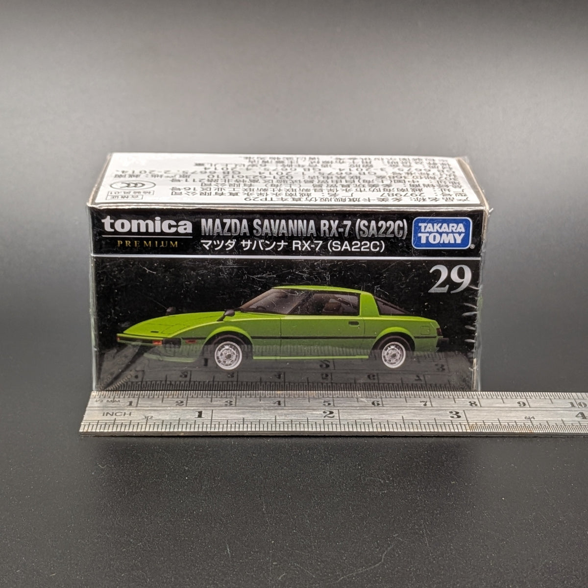 A boxed Tomica Premium Takara Toy Mazda Savanna RX-7 (SA22C) model car, shown in its original packaging with a clear plastic front and a ruler placed in front for scale on a dark gray background.