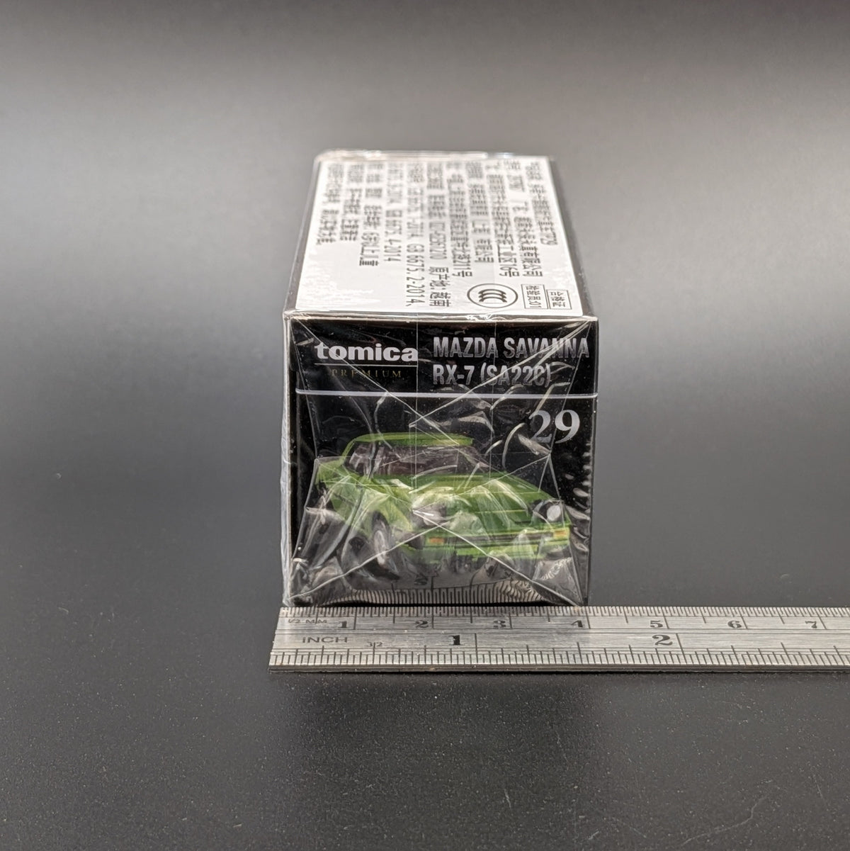 A sealed Tomica Mazda Savanna RX-7 toy car box, showing the green car printed on the front, with a metal ruler placed underneath to indicate scale, set against a dark gray background.
