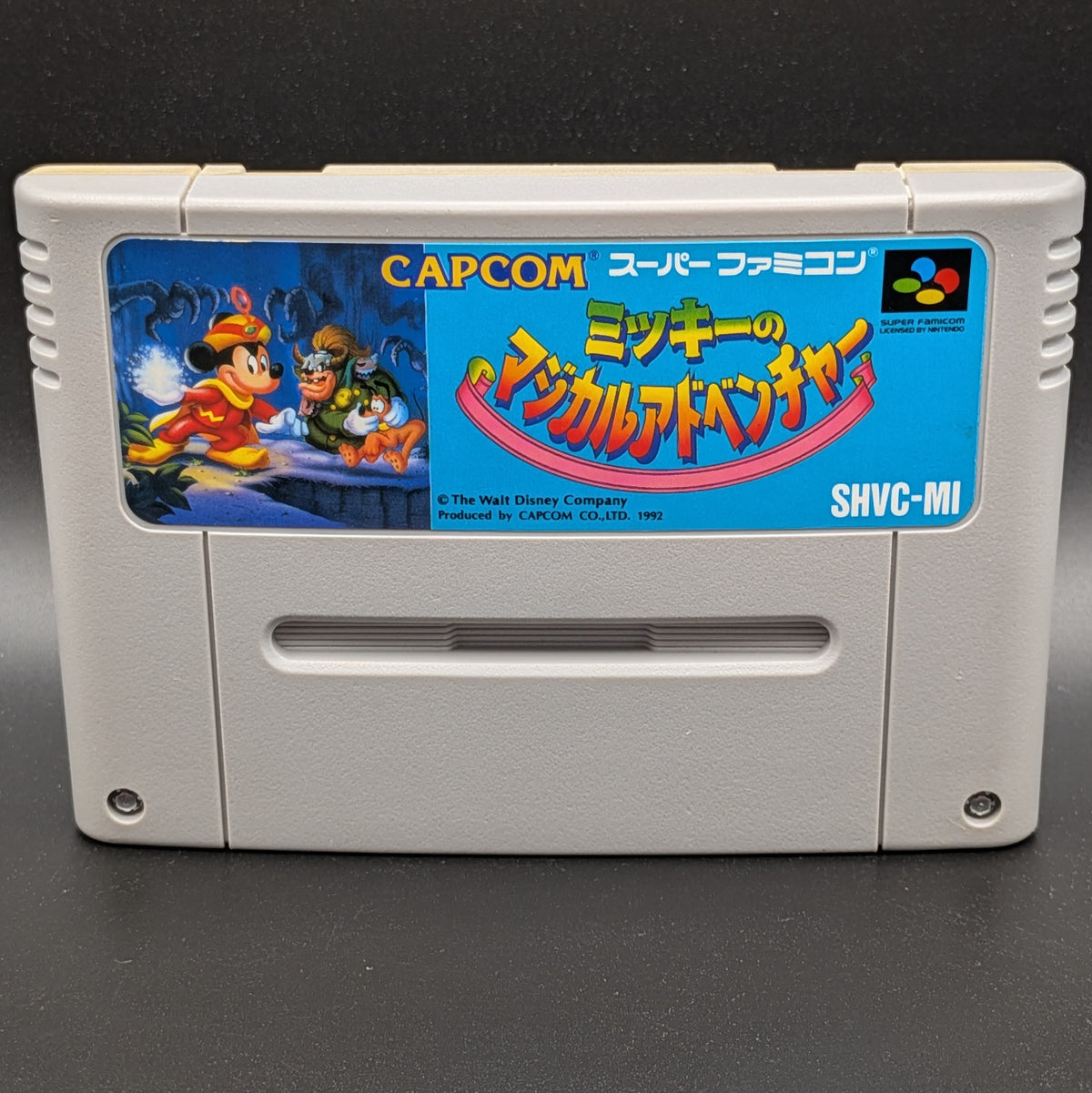 A close up of Mickey's Magical Adventure video game cartridge for Nintendo's Super Famicom (SFC) Entertainment System, shown standing up and facing forwards on a dark grey background.