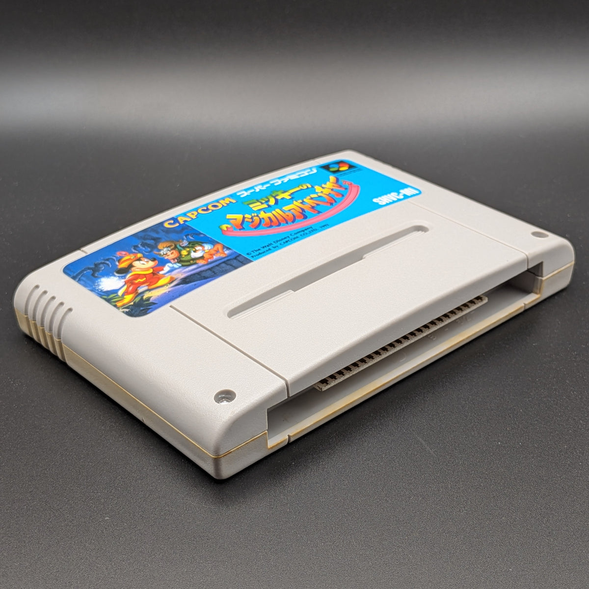 Mickey's Magical Adventure video game cartridge for Nintendo's Super Famicom (SFC) Entertainment System, shown laying on its back with the label facing upwards. Showing the left hand side and bottom slot. It is displayed on a dark grey background.