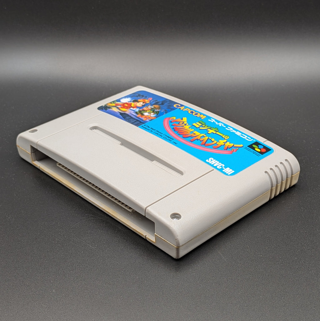 Mickey's Magical Adventure video game cartridge for Nintendo's Super Famicom (SFC) Entertainment System, shown laying on its back with the label facing upwards. Showing the right hand side and bottom slot. It is displayed on a dark grey background.