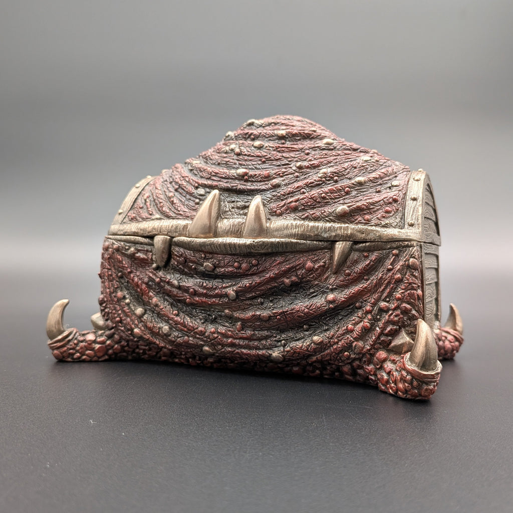 A fantasy miniature chest designed to look like a creature's scaly skin, featuring textured reddish-brown scales, spikes, and claw-like feet on the sides, set against a neutral gray background.
