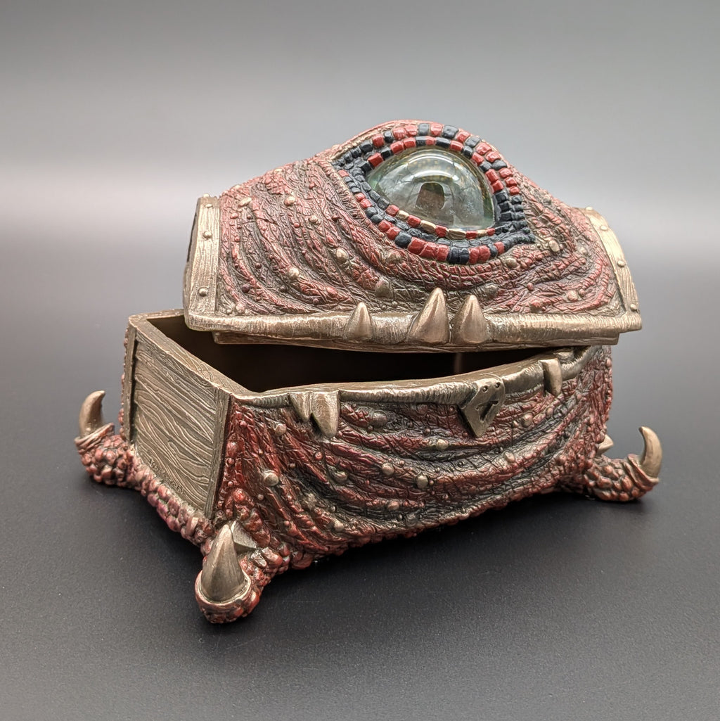 A fantasy creature eye chest or box with a textured, reptilian lid featuring a prominent green eye and red and gold bead accents, standing on clawed feet on a dark gray background.