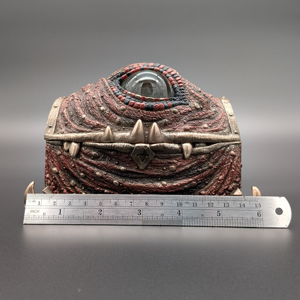 A fantasy one-eyed treasure chest figurine with a textured red and brown body, metallic bronze straps, and a clear glass eye, shown with a metal ruler in front on a dark gray background.