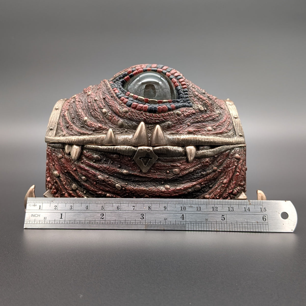 A fantasy one-eyed treasure chest figurine with a textured red and brown body, metallic bronze straps, and a clear glass eye, shown with a metal ruler in front on a dark gray background.