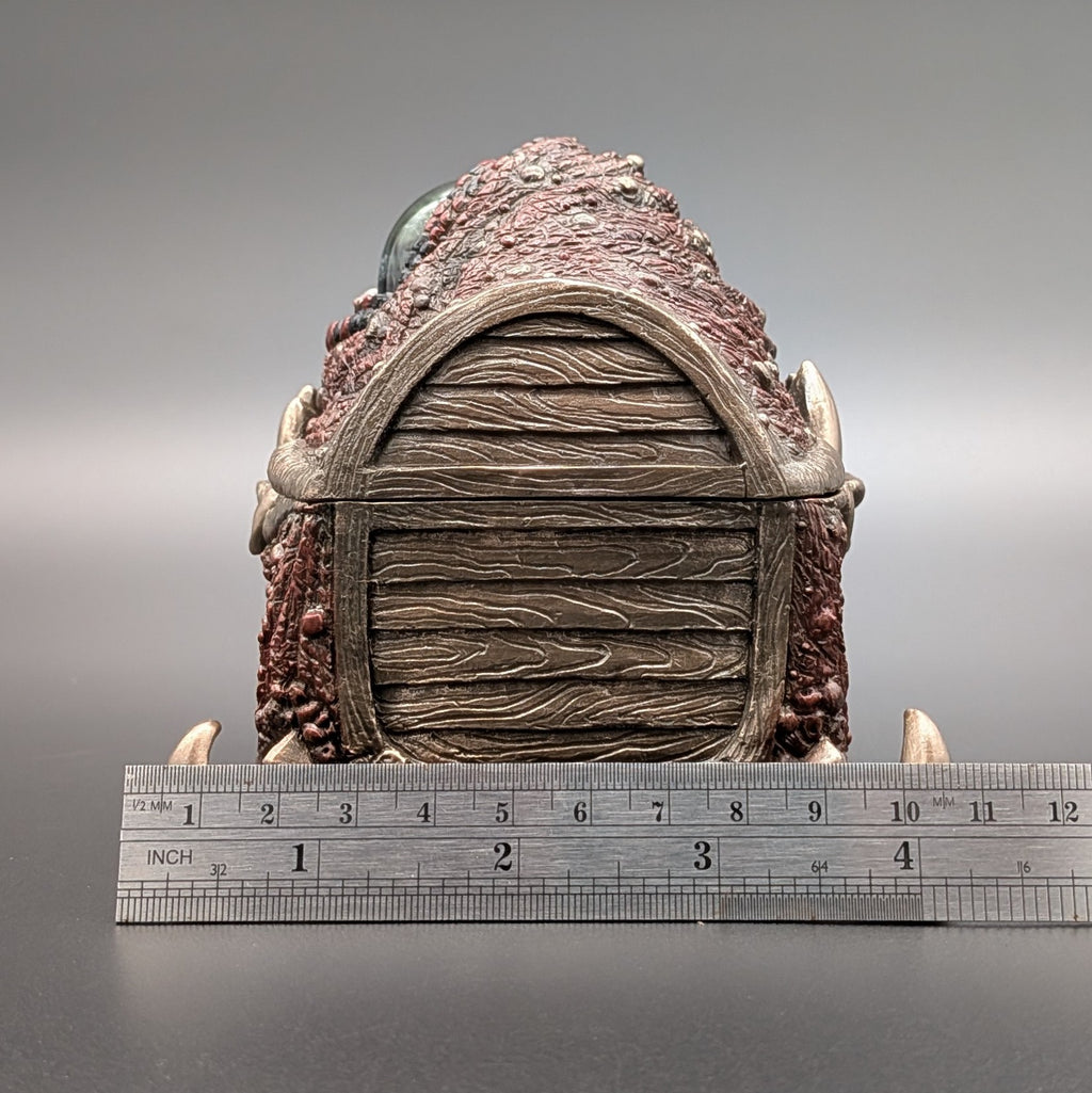 A fantasy creature chest collectible piece with a textured brown and reddish exterior, a wood-grain patterned side, and horn-like protrusions, shown upright with a metal ruler below for scale on a gray studio background.