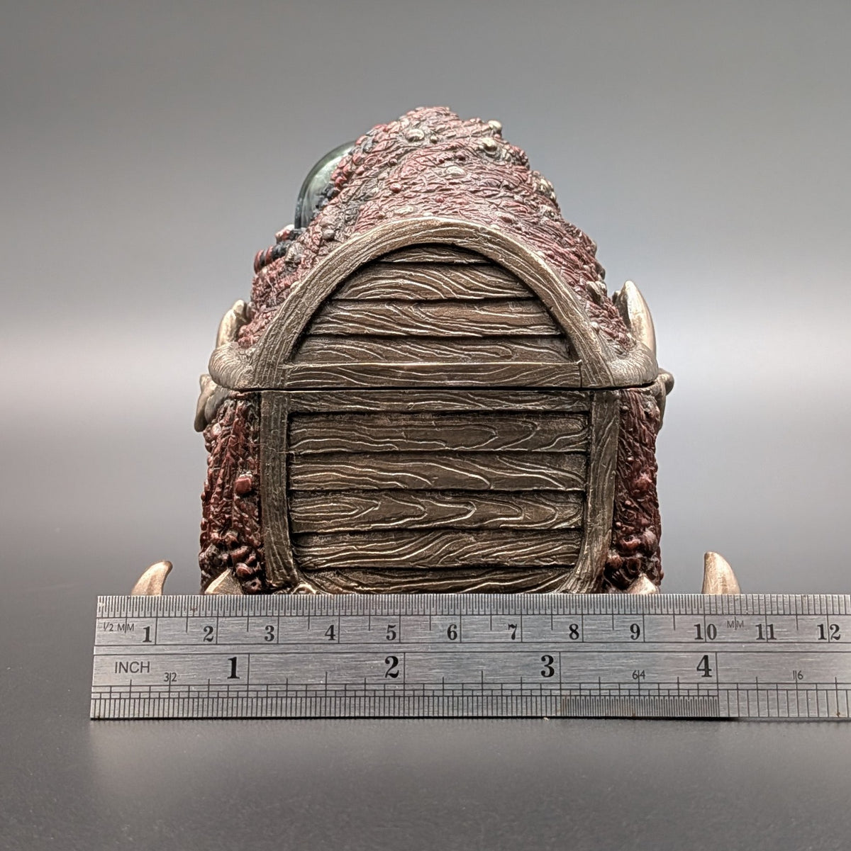 A fantasy creature chest collectible piece with a textured brown and reddish exterior, a wood-grain patterned side, and horn-like protrusions, shown upright with a metal ruler below for scale on a gray studio background.