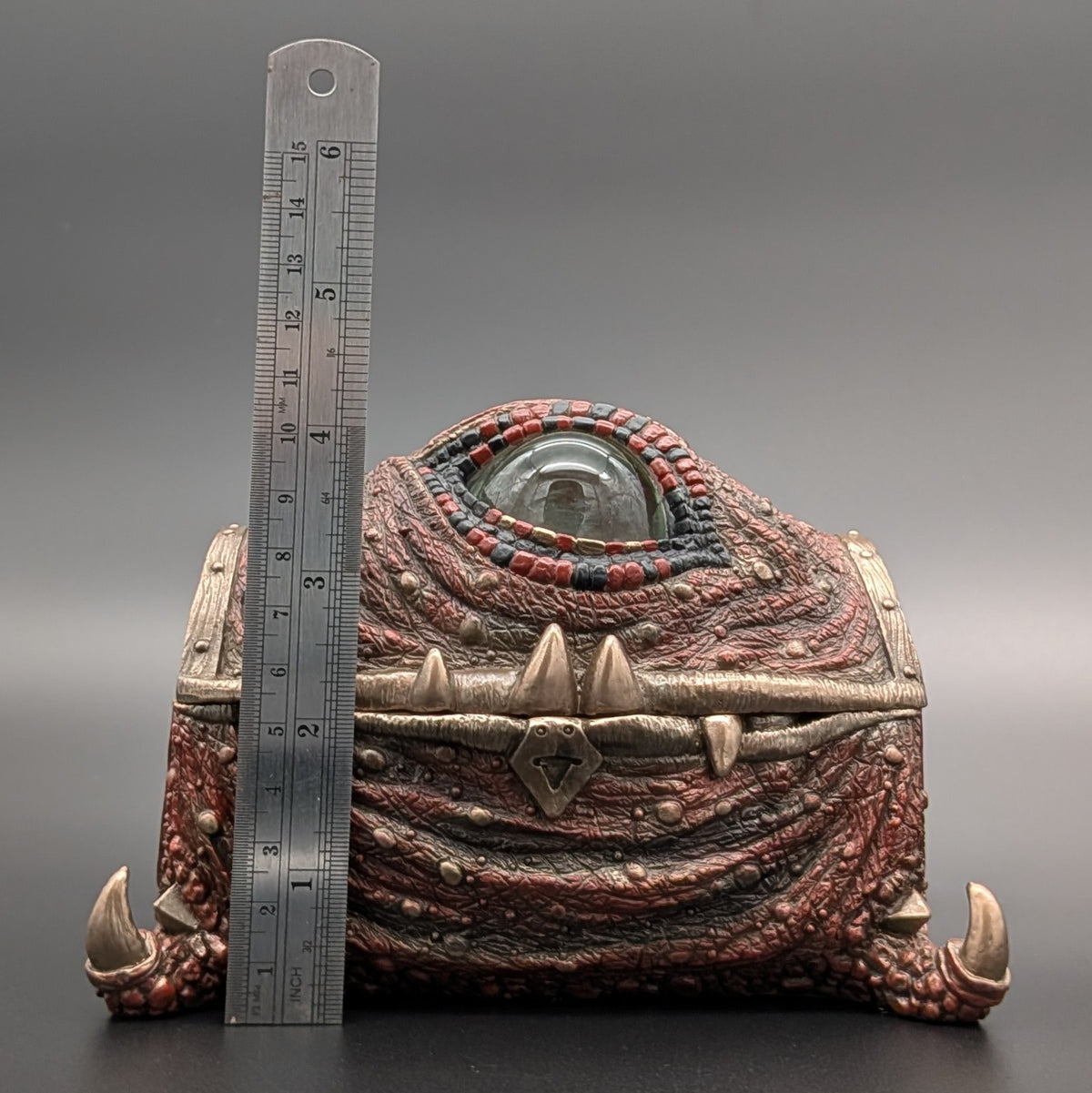 A fantasy one-eyed monster chest with textured reddish-brown scales, metallic bronze accents, and clawed feet, shown next to a metal ruler for scale on a gray background.