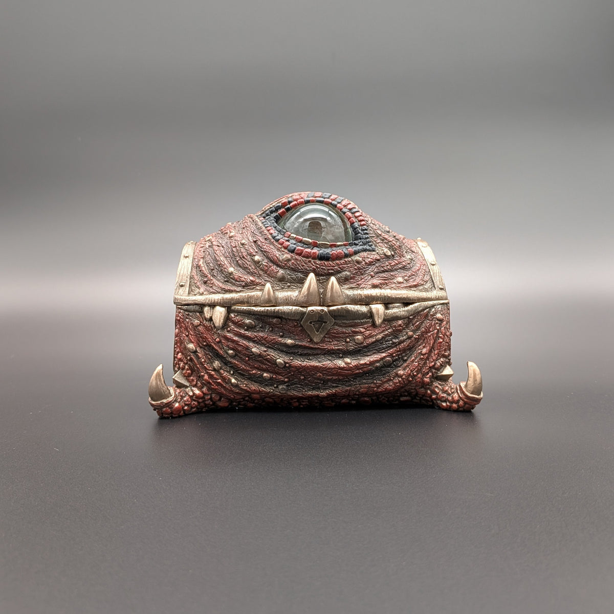 A fantasy one-eyed treasure chest figurine with textured red and brown drapery, spikes, and a single prominent eye on top, set against a dark gray gradient background.