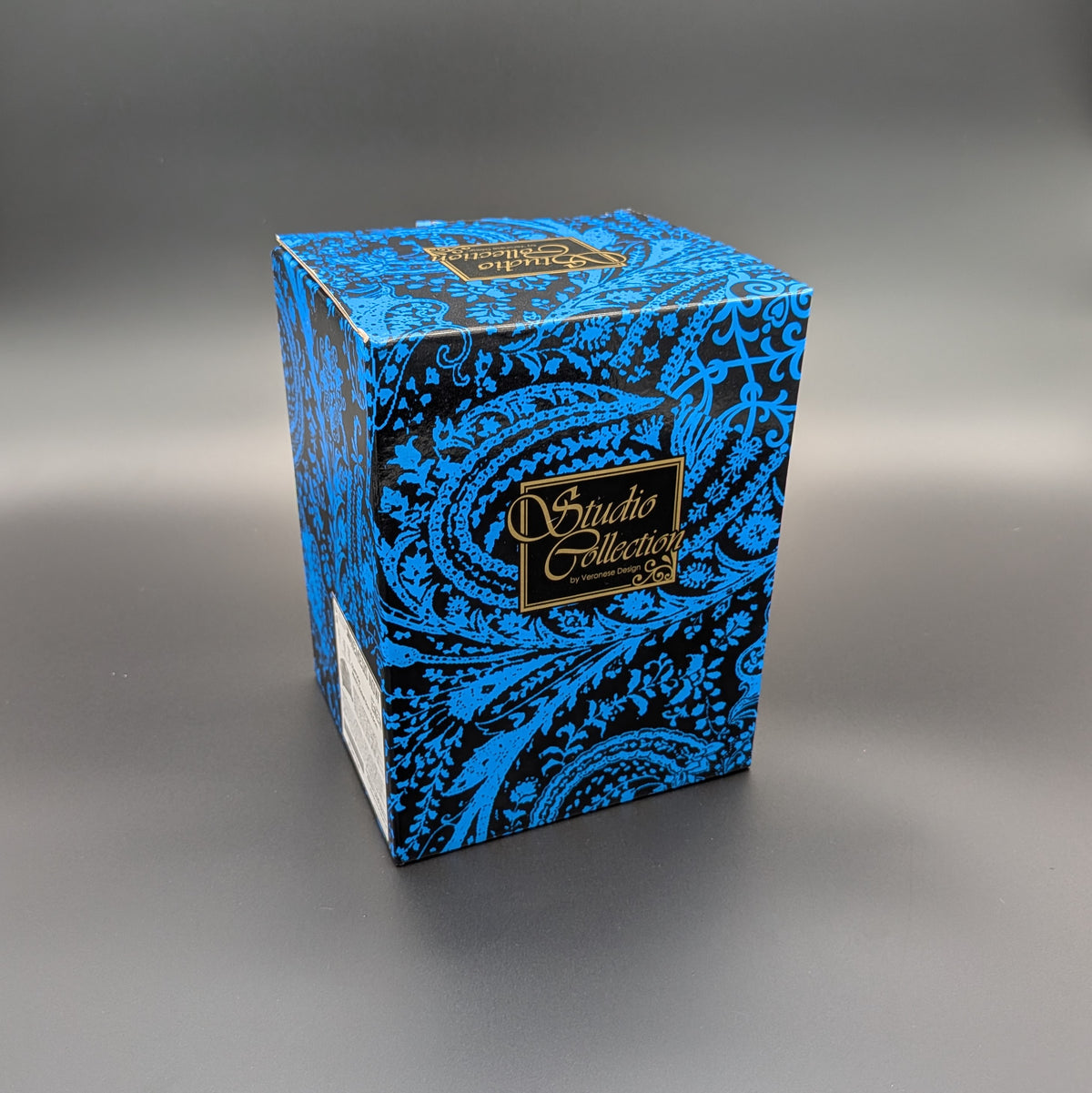 A square product box with a blue and black paisley pattern and a gold 'Studio Collection' logo, photographed on a dark gray gradient background.