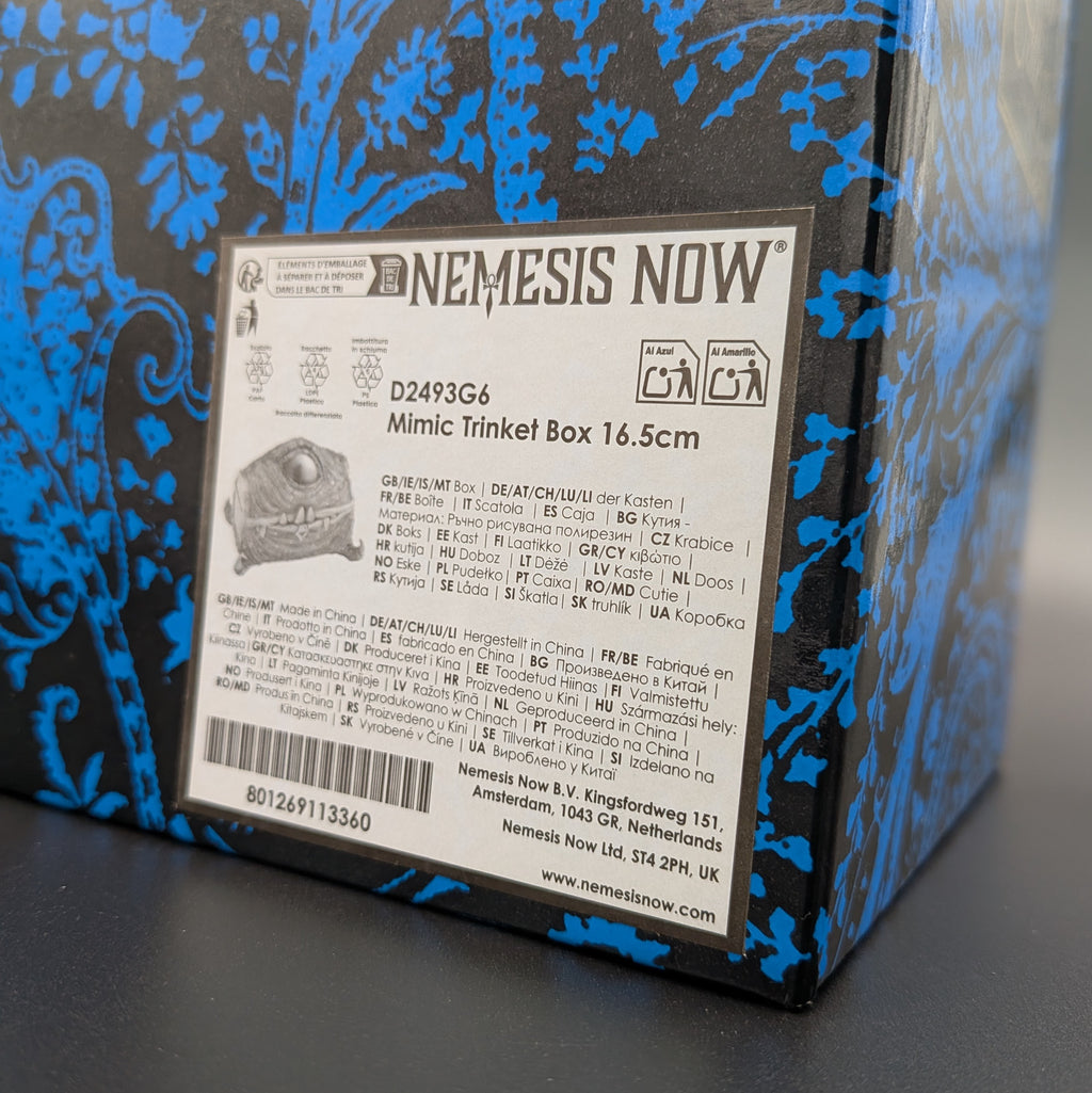 A close-up of a Nemesis Now Mimic Trinket Box, showing the back of the packaging with a detailed product label and the brand name 'NEMESIS NOW' printed on the side.